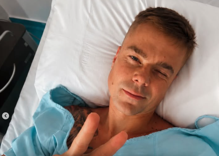 Ein Mann liegt im Krankenhausbett und zeigt ein Peace-Zeichen mit der Hand.