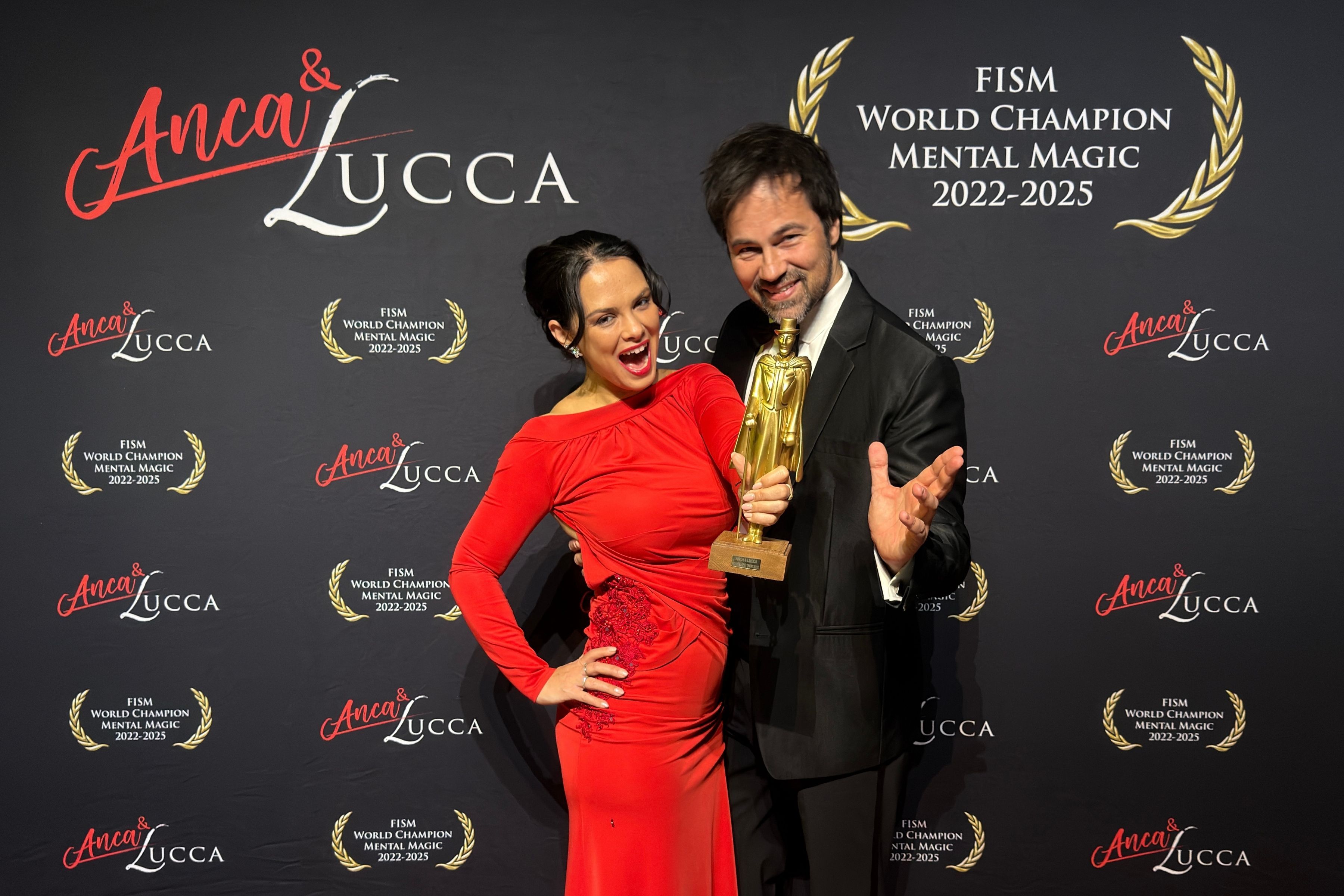 Anca und Lucca posieren mit einem goldenen Preis vor einer Wand mit ihrem Namen und Titeln.