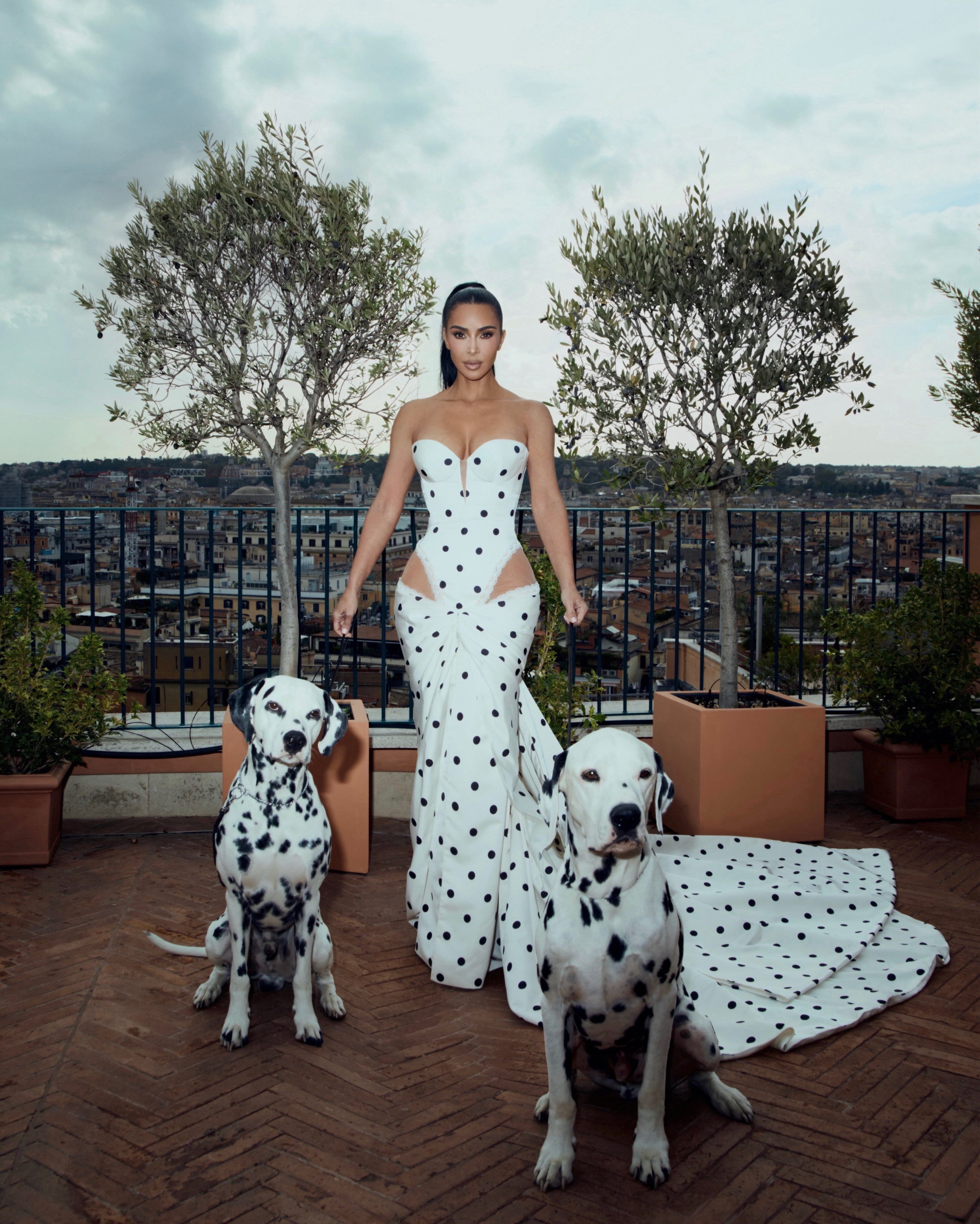Frau im weißen, gepunkteten Kleid steht mit zwei Dalmatinern auf einer Terrasse mit Stadtblick.