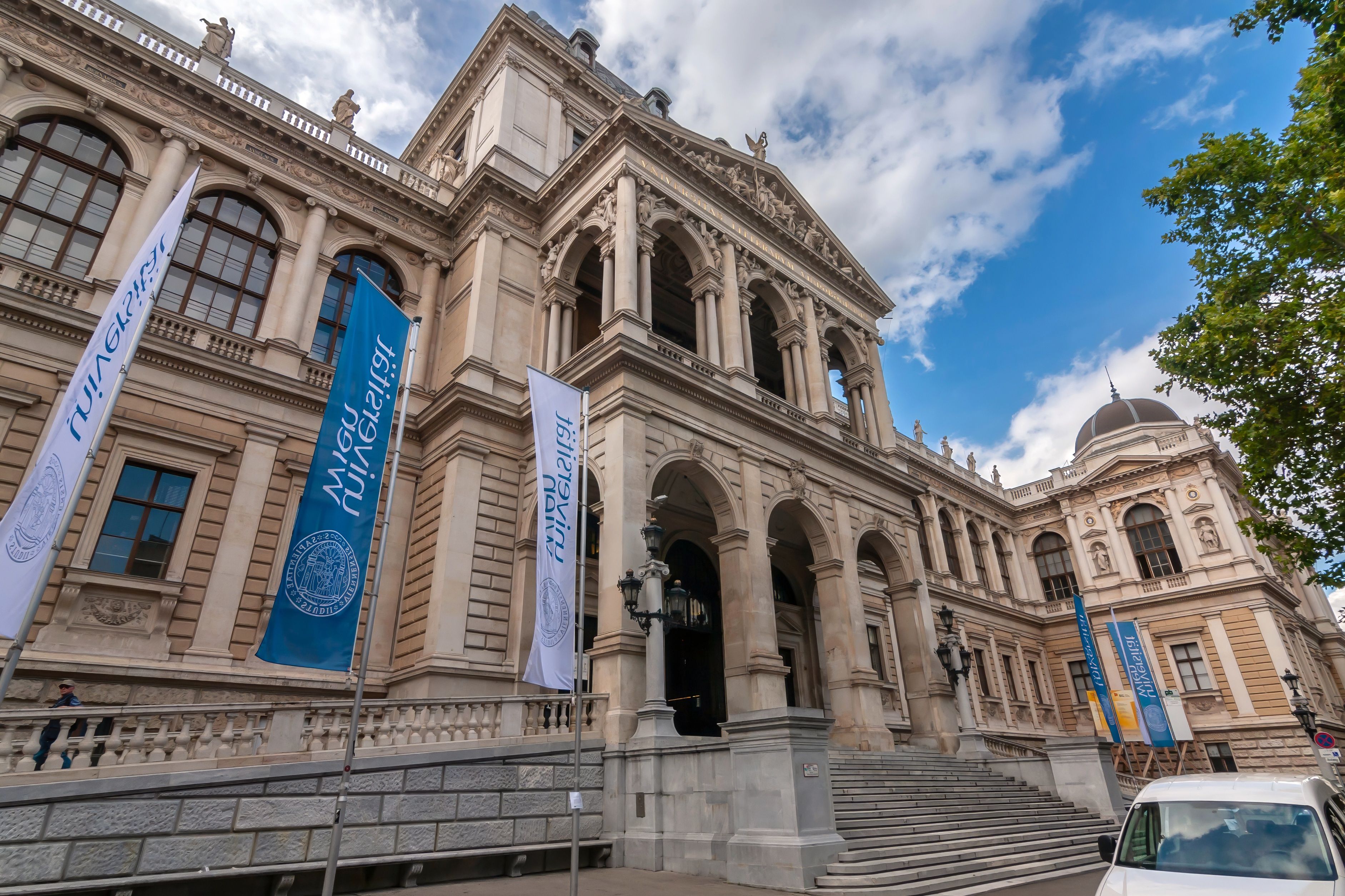 Das Hauptgebäude der Universität Wien mit Fahnen und breiter Freitreppe bei blauem Himmel.