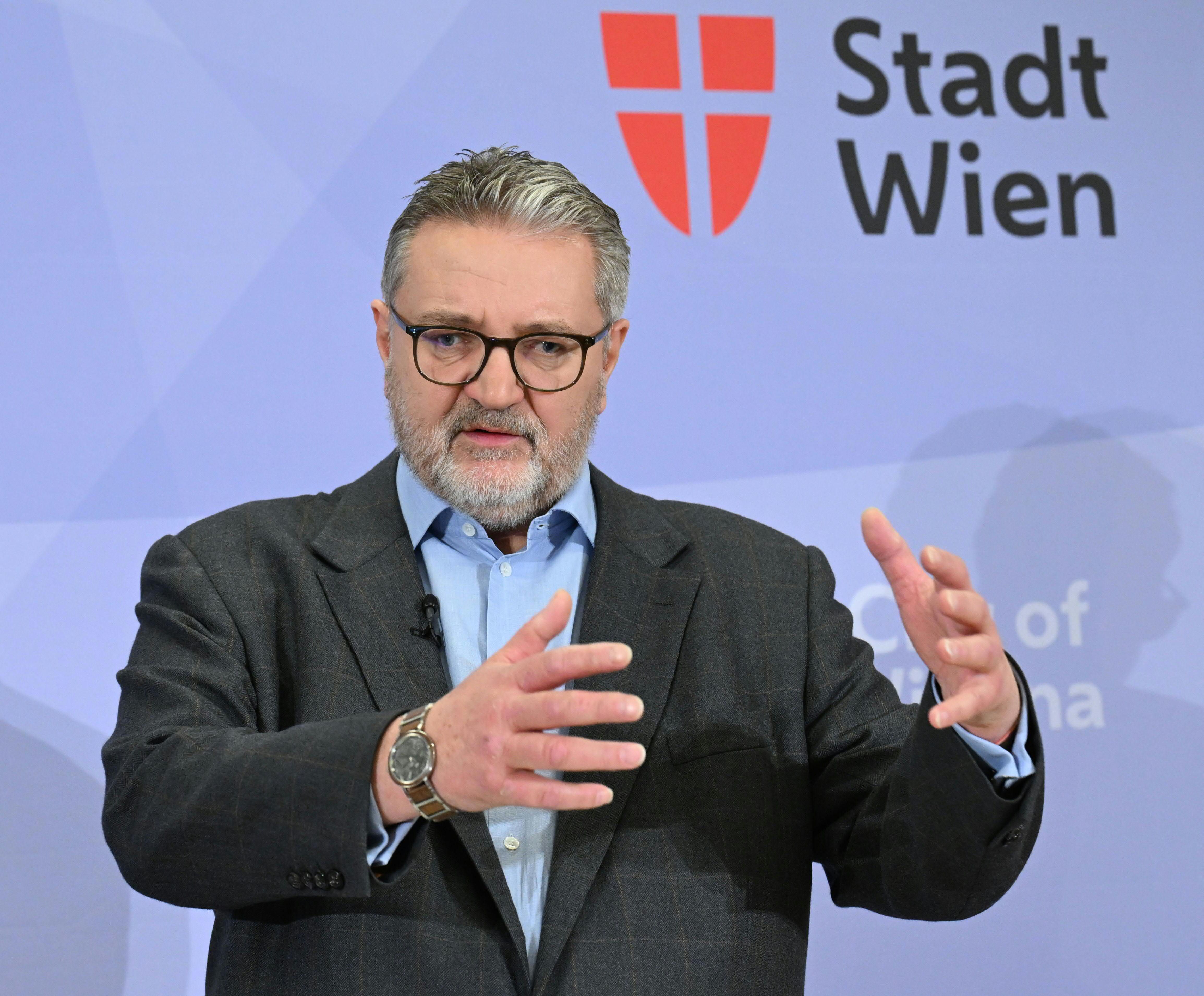 Ein Mann mit Brille und Bart spricht gestikulierend vor einem Logo der Stadt Wien.