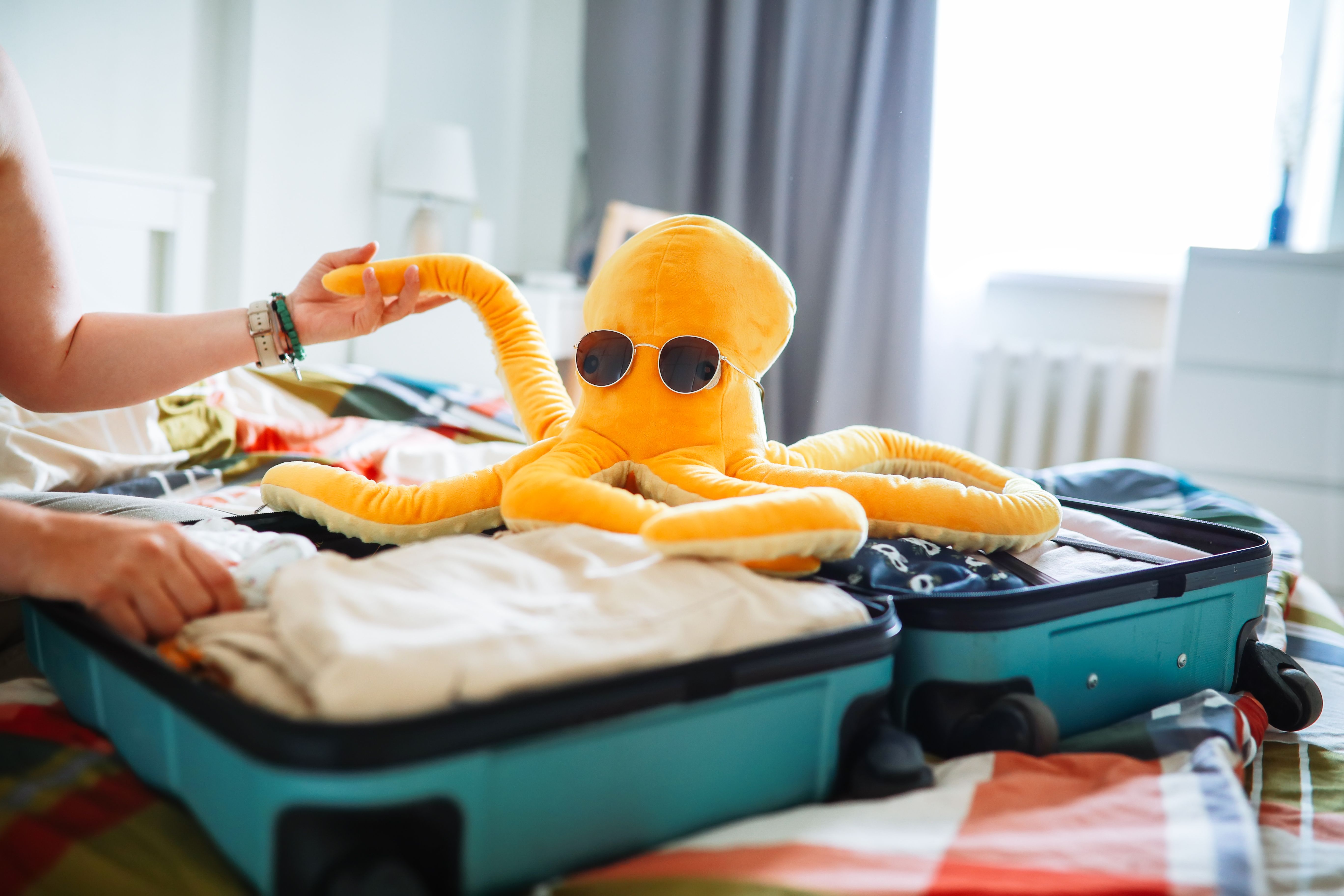 Gelbes Plüsch-Oktopus mit Sonnenbrille sitzt im gepackten Koffer auf einem Bett.