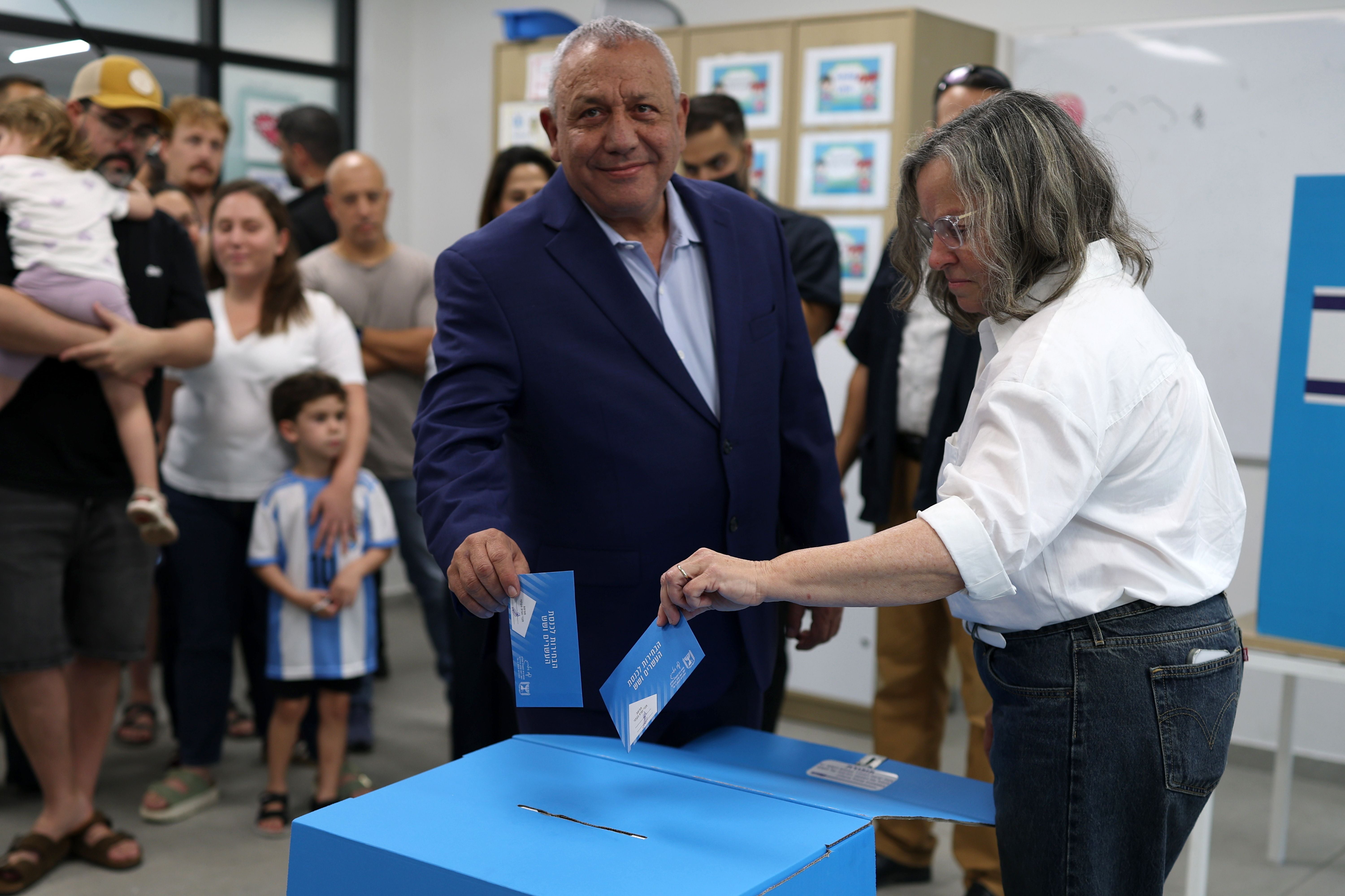 Ein Mann im Anzug und eine Frau werfen Wahlzettel in eine Wahlurne, Menschen stehen im Hintergrund.