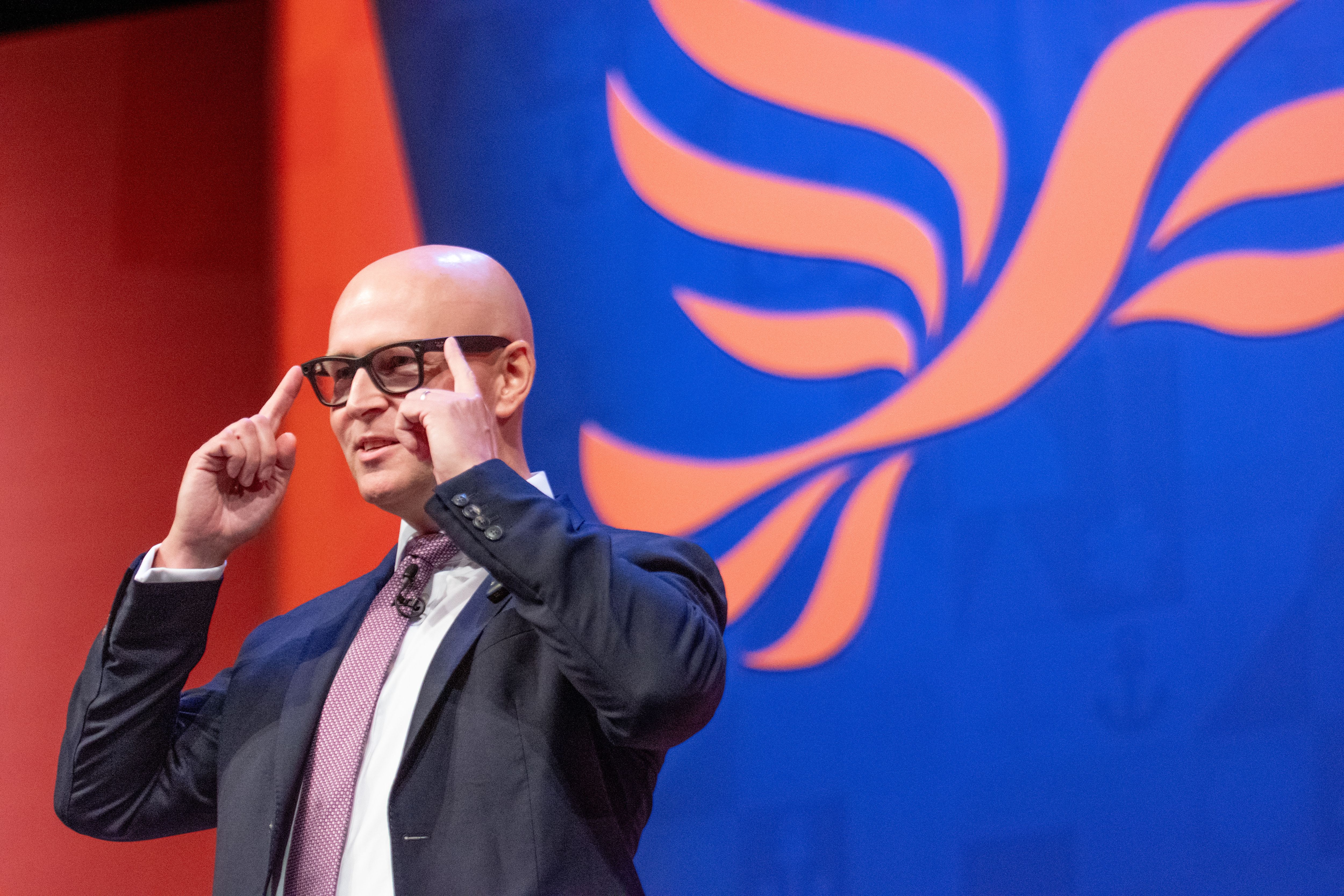 Ein Mann mit Brille spricht auf einer Bühne vor einem blauen Hintergrund mit orangenem Flügelmotiv.