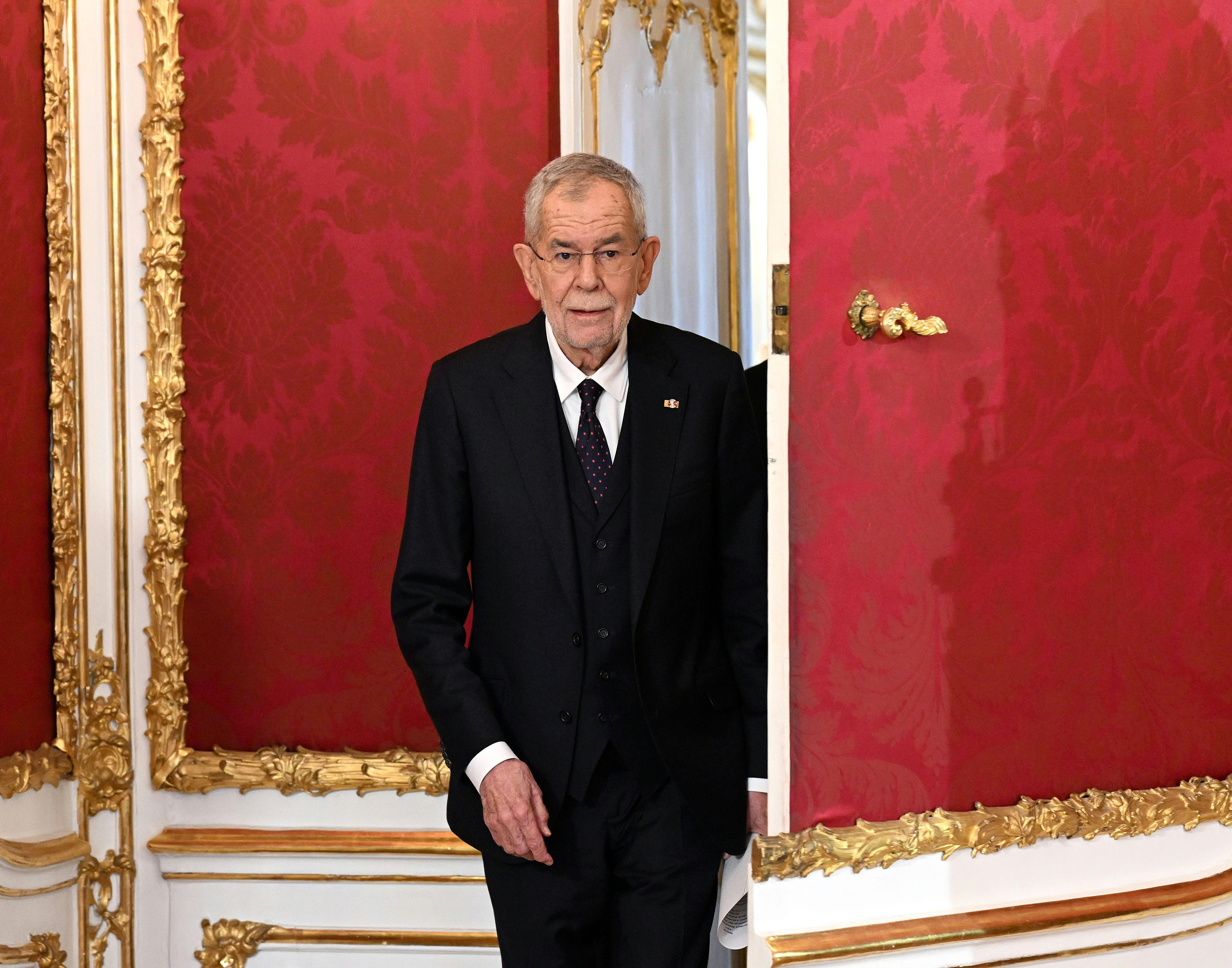 Ein älterer Mann im Anzug tritt durch eine reich verzierte Tür in einem prunkvollen, roten Saal.
