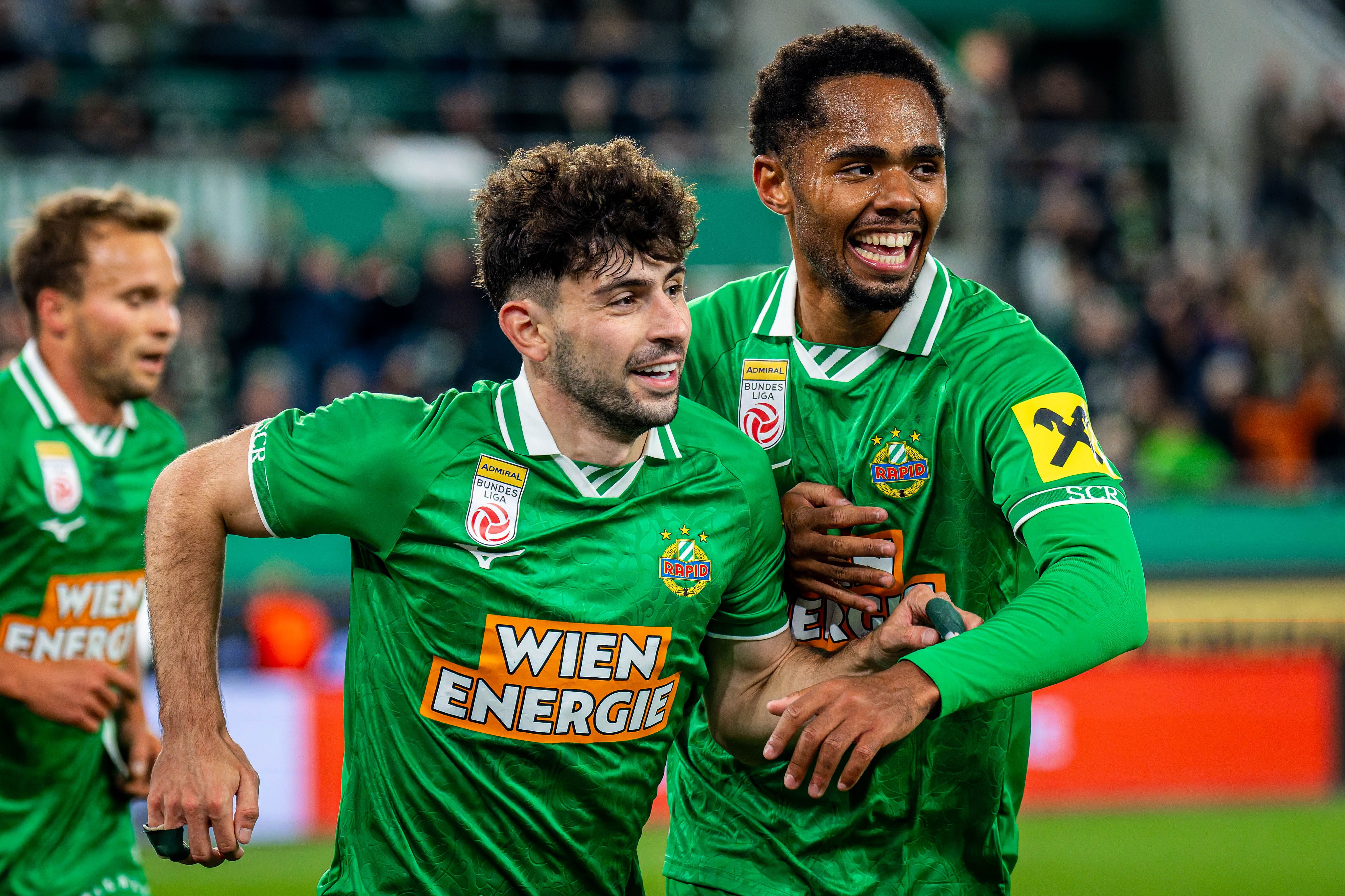 Zwei Rapid Wien Fußballspieler jubeln im grünen Trikot auf dem Spielfeld.