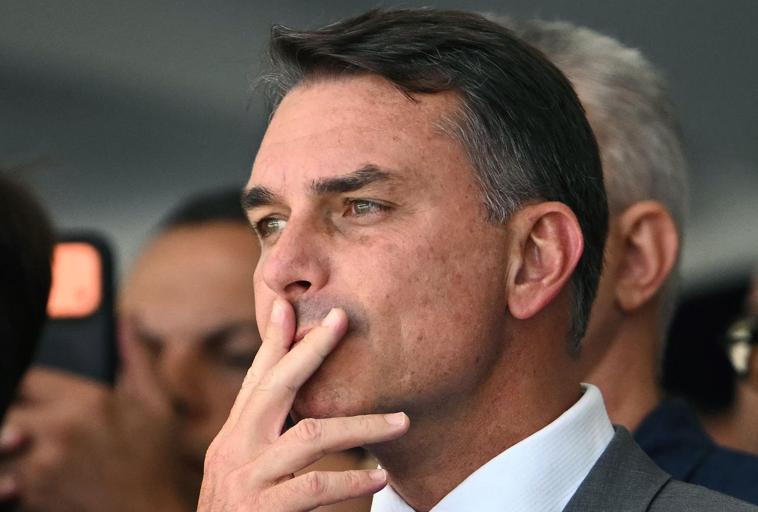 Ein Mann im Anzug hält nachdenklich seine Finger ans Gesicht, im Hintergrund weitere Personen.