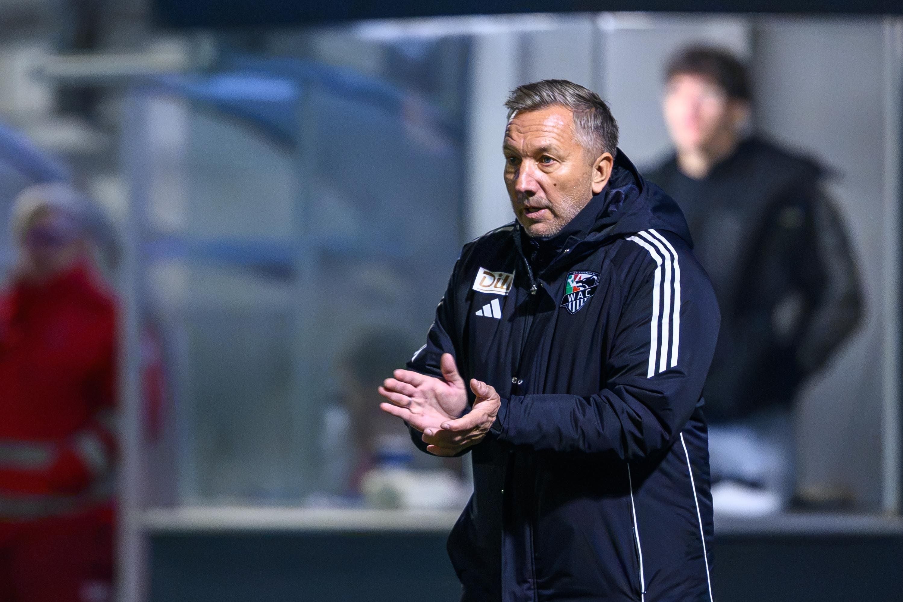 Manfred Schmid klatscht in dunkler Trainingsjacke mit dem Logo des Wolfsberger AC am Spielfeldrand aufmerksam in die Hände.