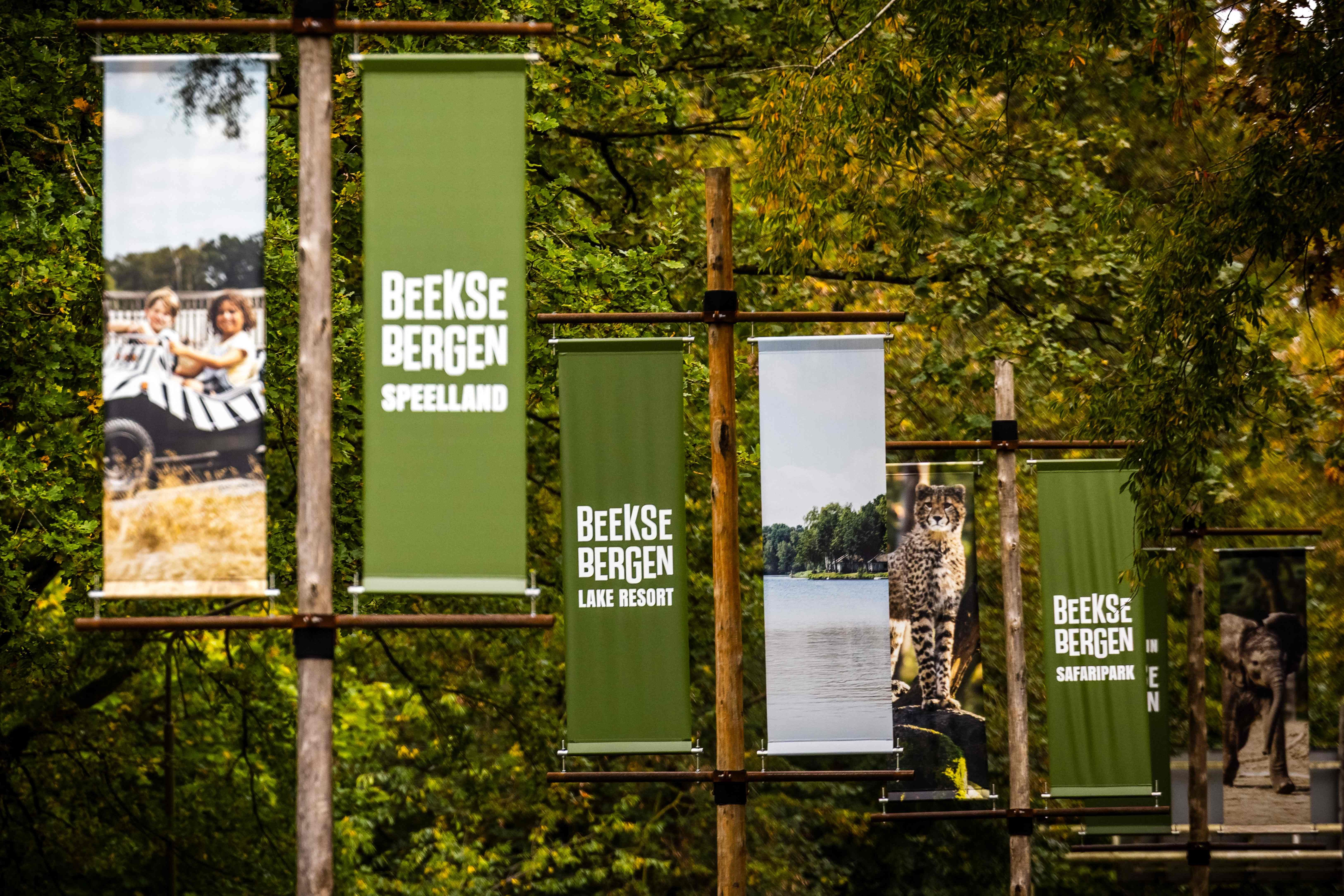 Mehrere grüne Banner mit "Beekse Bergen" am Eingang eines Safariparks, von Bäumen umgeben.