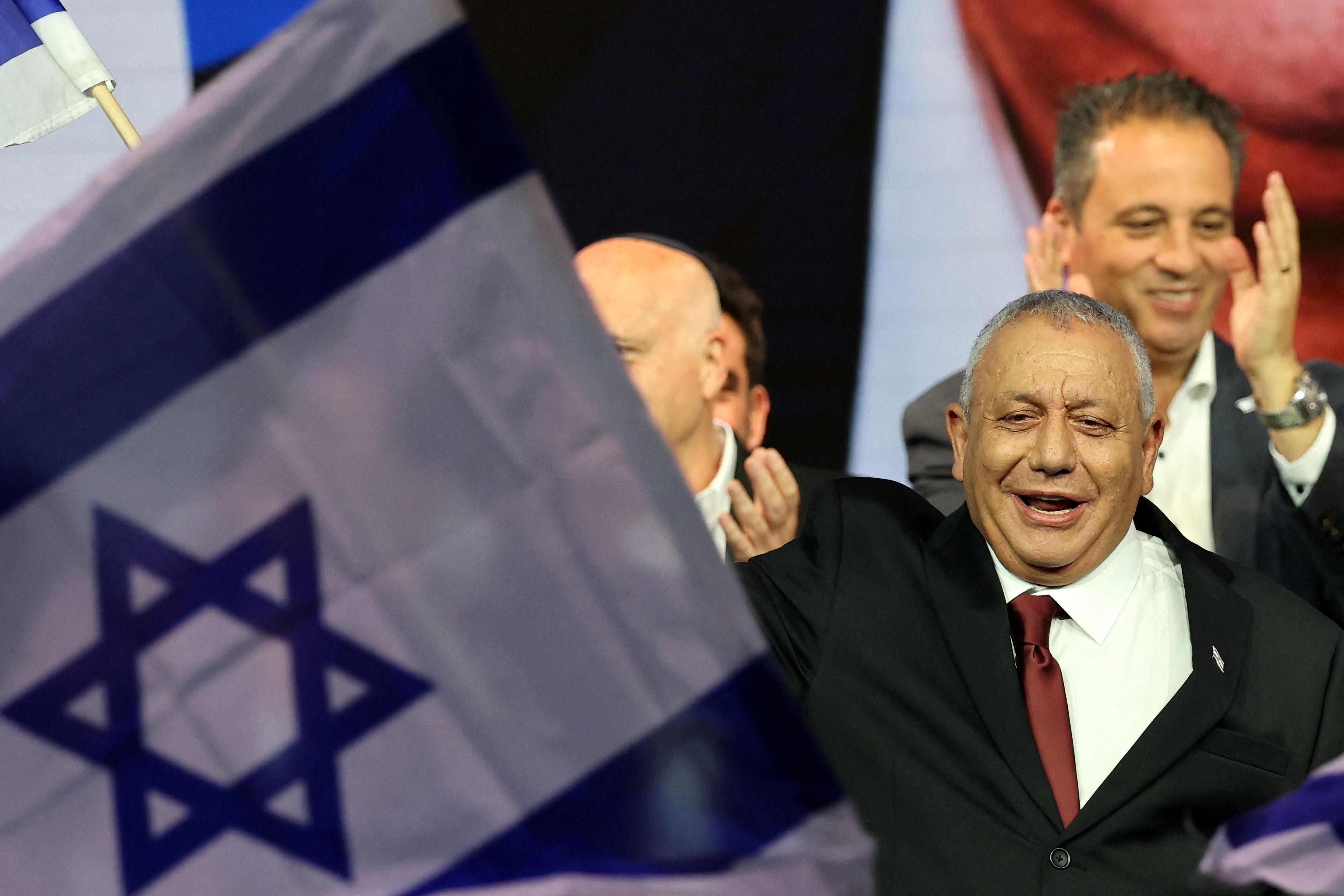 Ein lachender Mann im Anzug winkt, eine große israelische Flagge im Vordergrund.