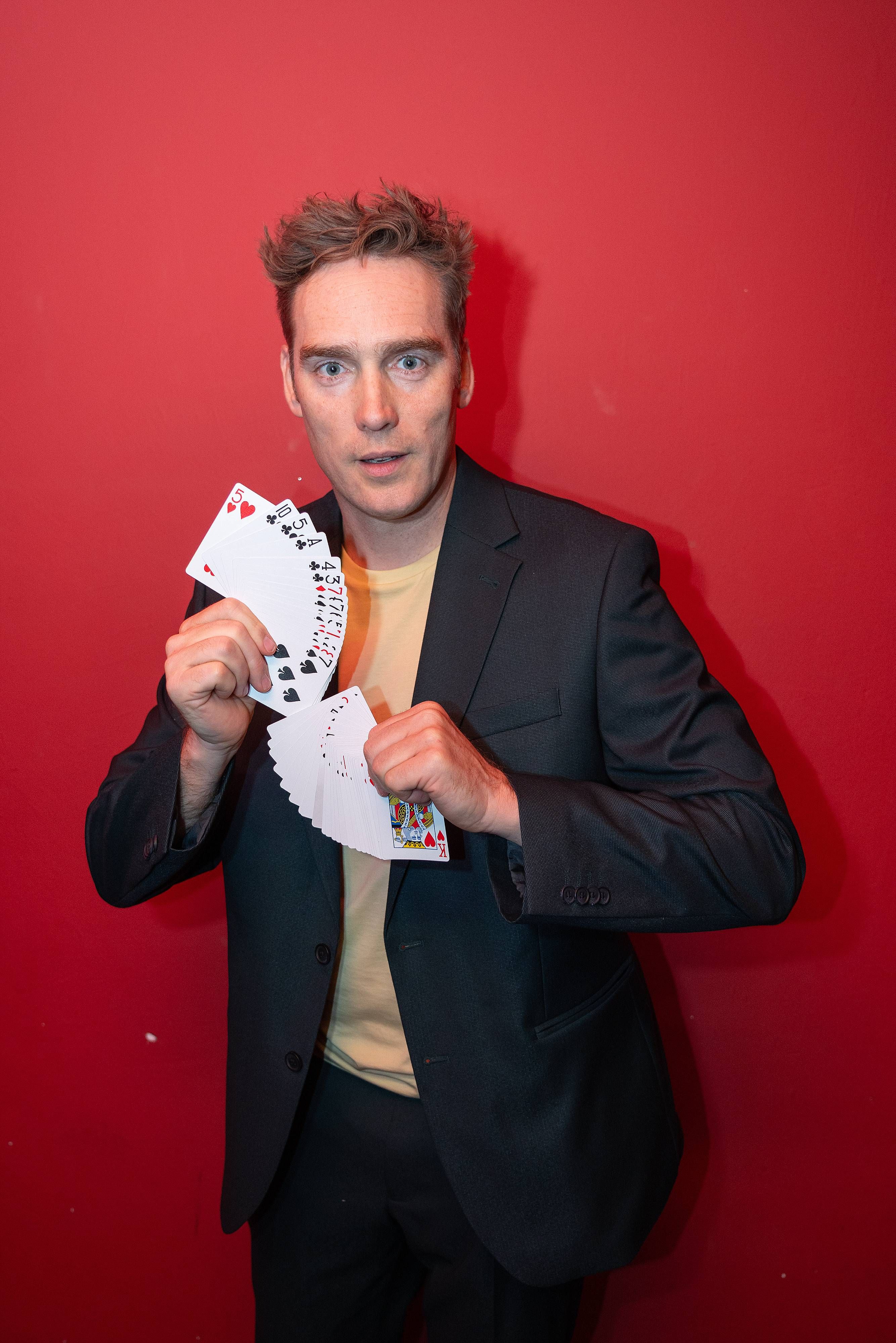 Ein Mann im Anzug hält aufgefächerte Spielkarten vor rotem Hintergrund.
