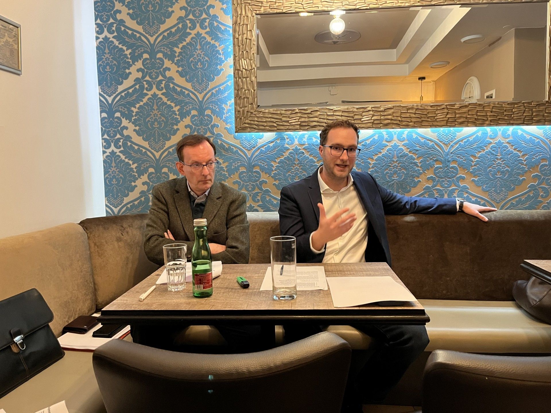 Zwei Männer sitzen in einem Café an einem Tisch mit Wasserflaschen und Papieren, einer spricht.