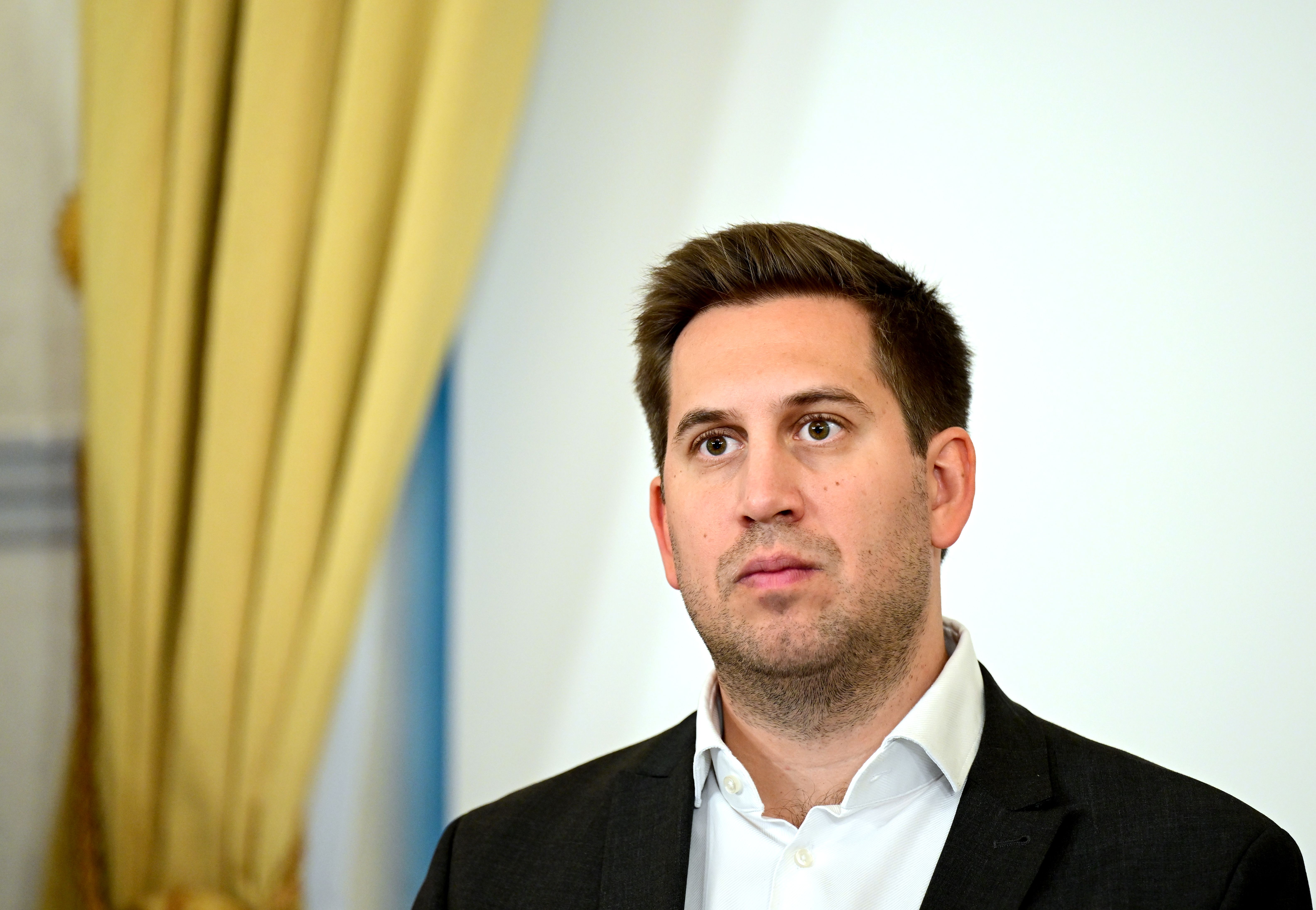 Ein Mann im Anzug steht vor einem hellen Hintergrund mit gelbem Vorhang.
