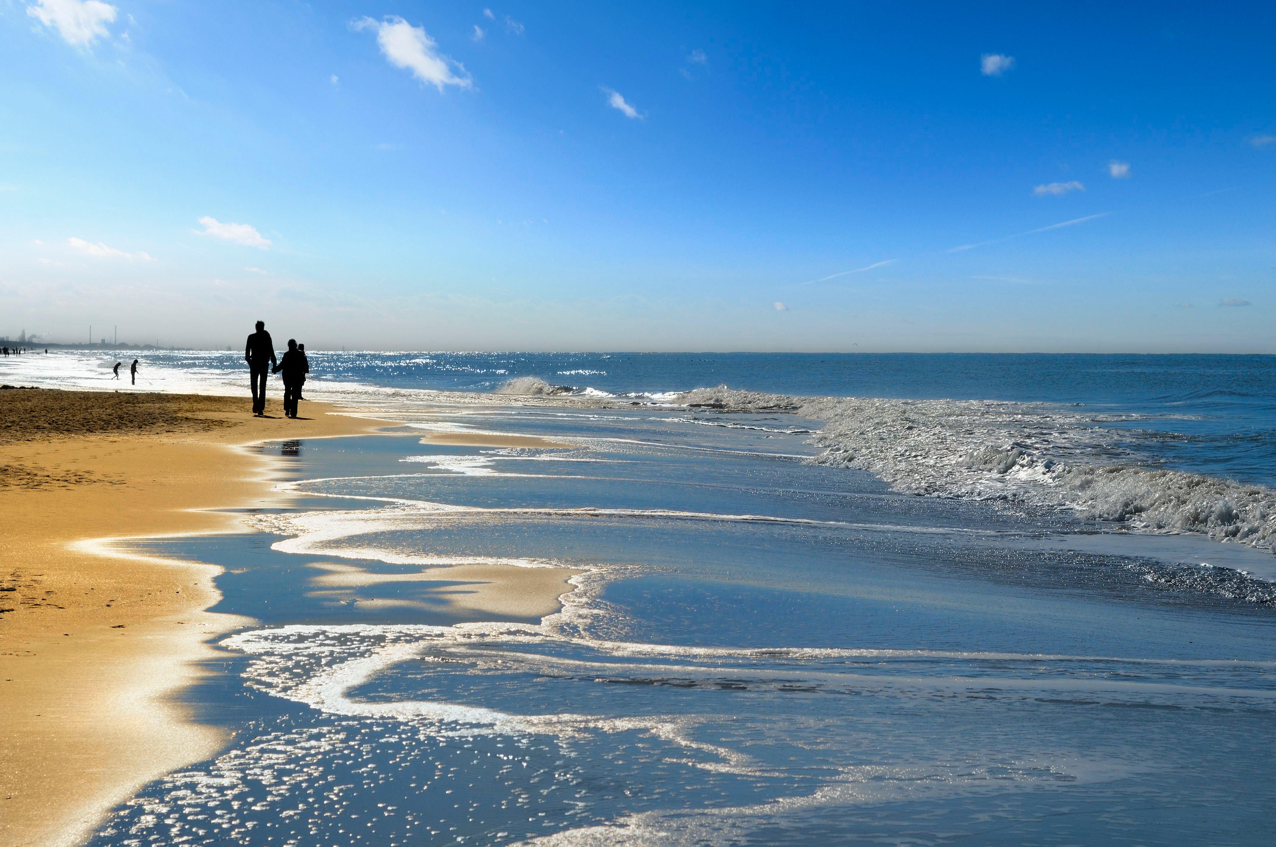 Zwei Menschen gehen am sonnigen Strand der Nordsee, Wellen spiegeln das Sonnenlicht wider.