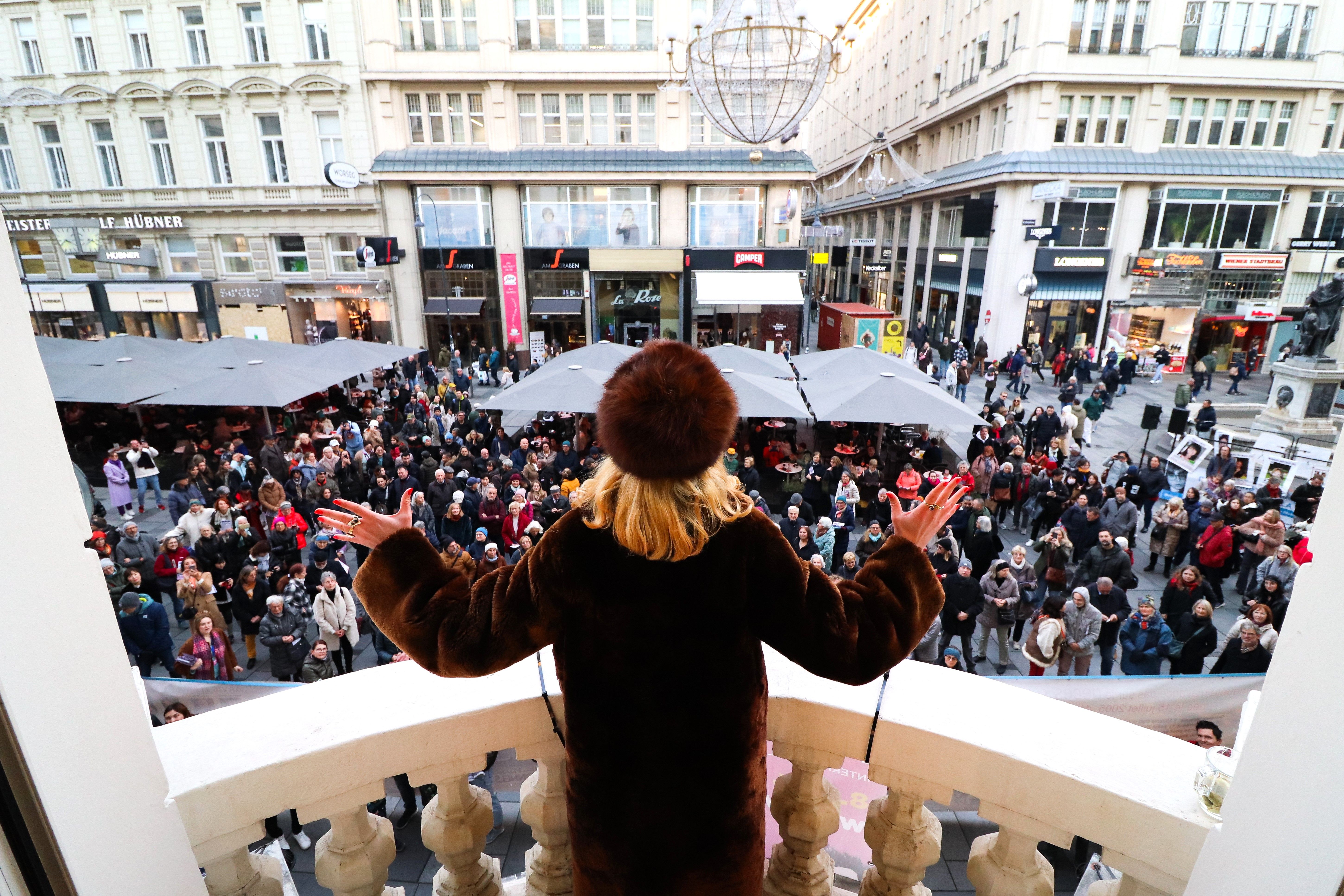 Eine Frau im Pelzmantel steht auf einem Balkon und spricht zu einer Menschenmenge auf der Straße.