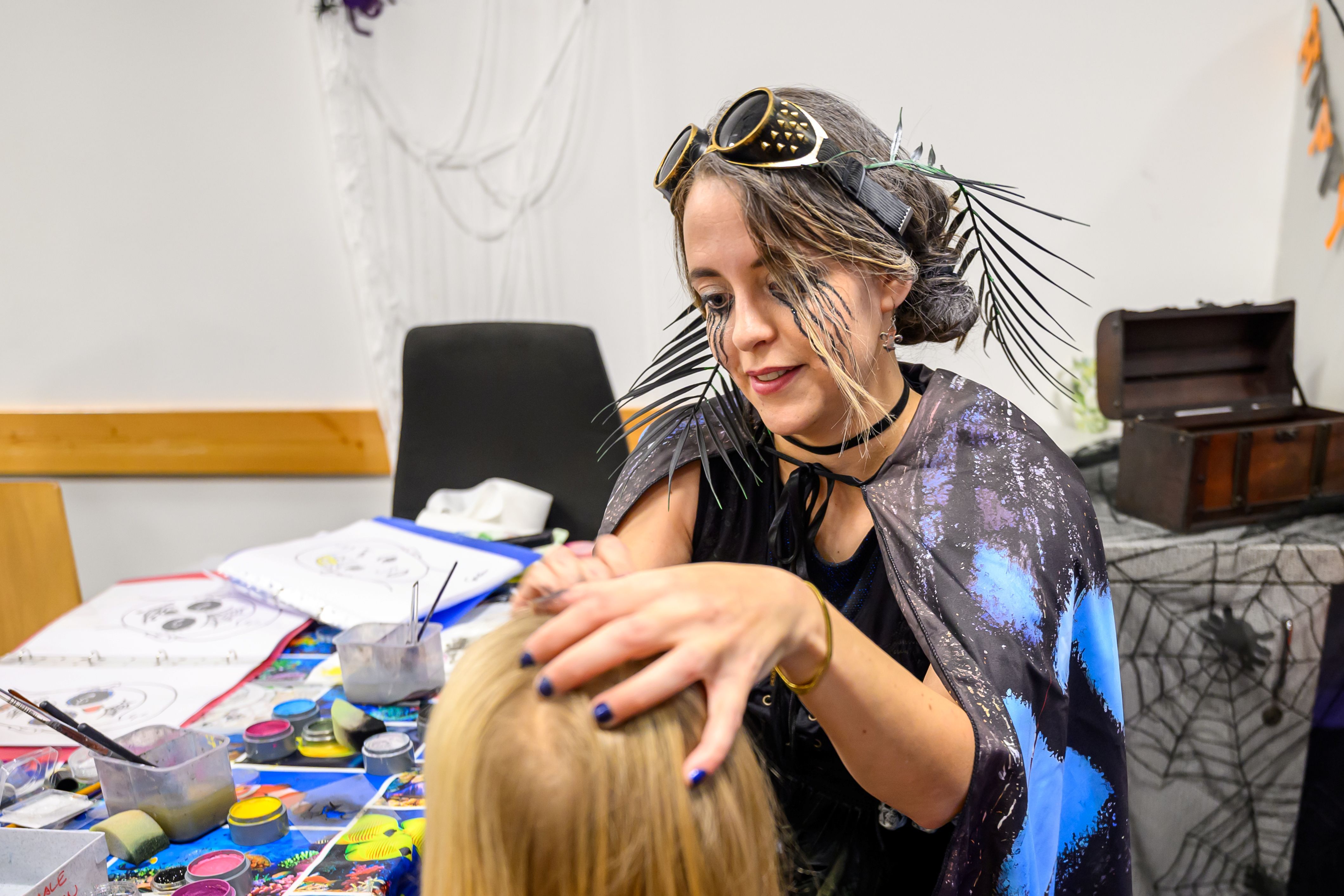 Frau im Halloween-Kostüm schminkt ein Kind, Tisch mit Schminkutensilien im Vordergrund.