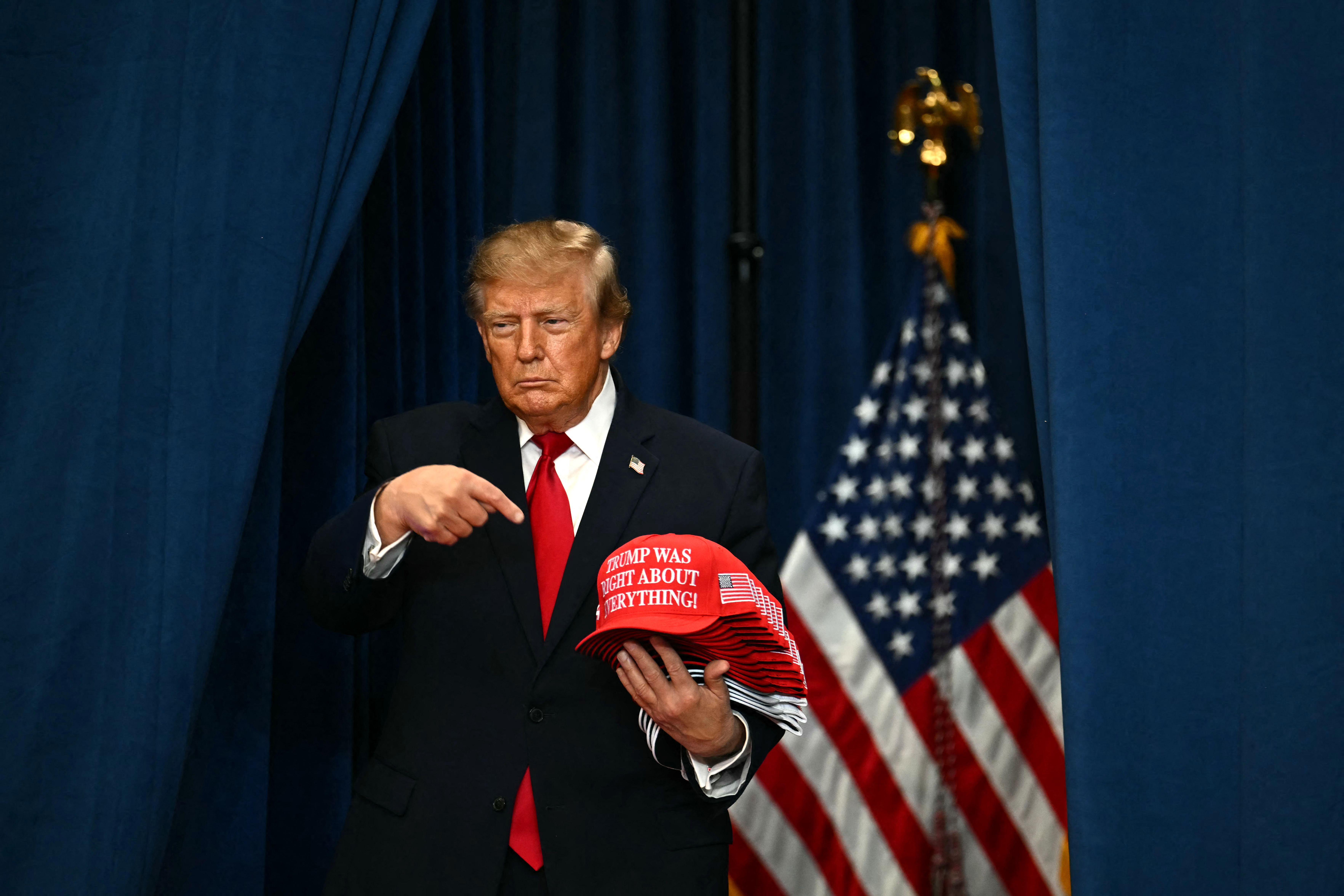 Donald Trump hält rote Kappen vor einer amerikanischen Flagge und steht vor blauen Vorhängen.