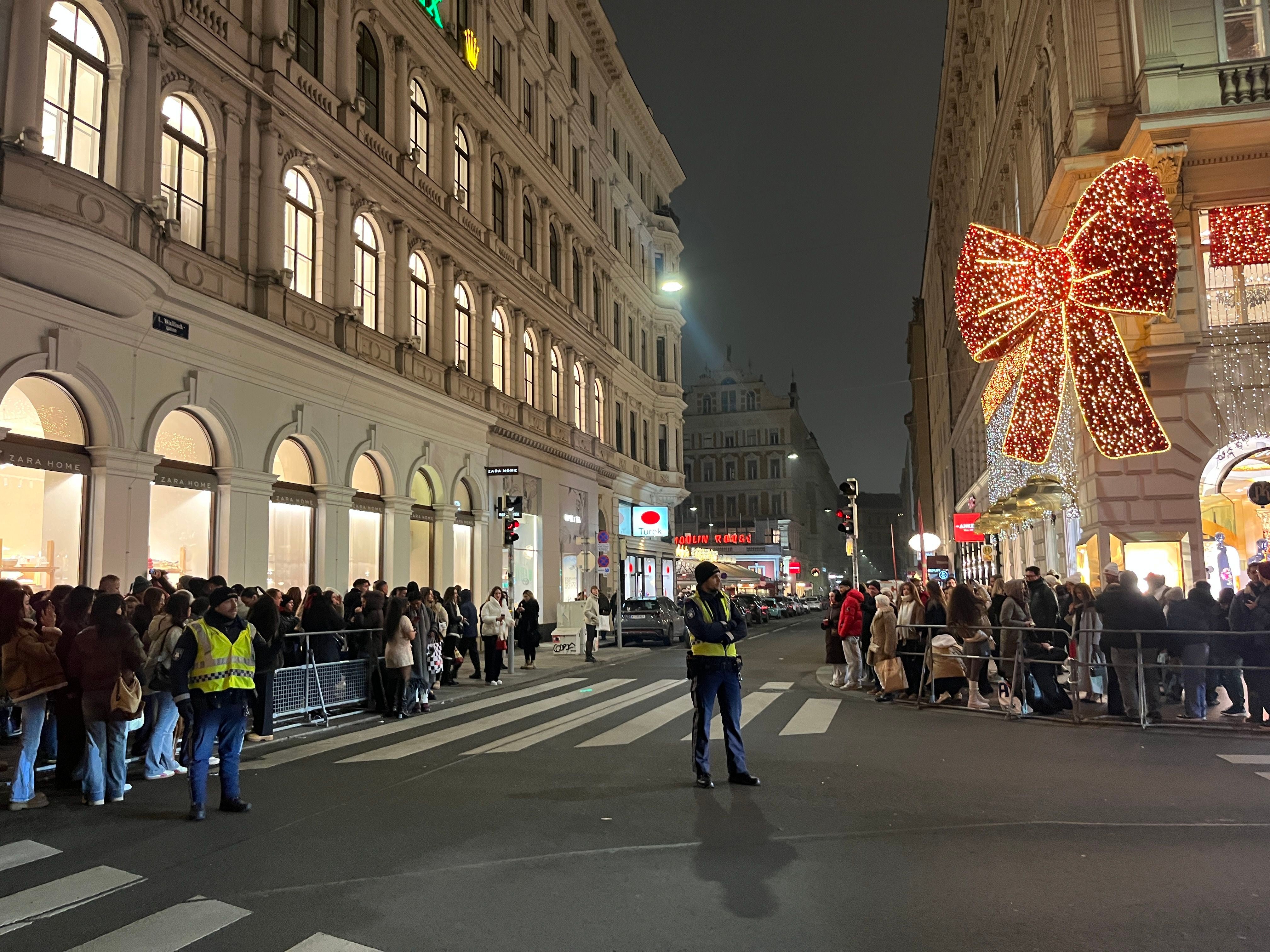 Menschen stehen nachts in einer Schlange vor Geschäften, große leuchtende Schleife an der Fassade.