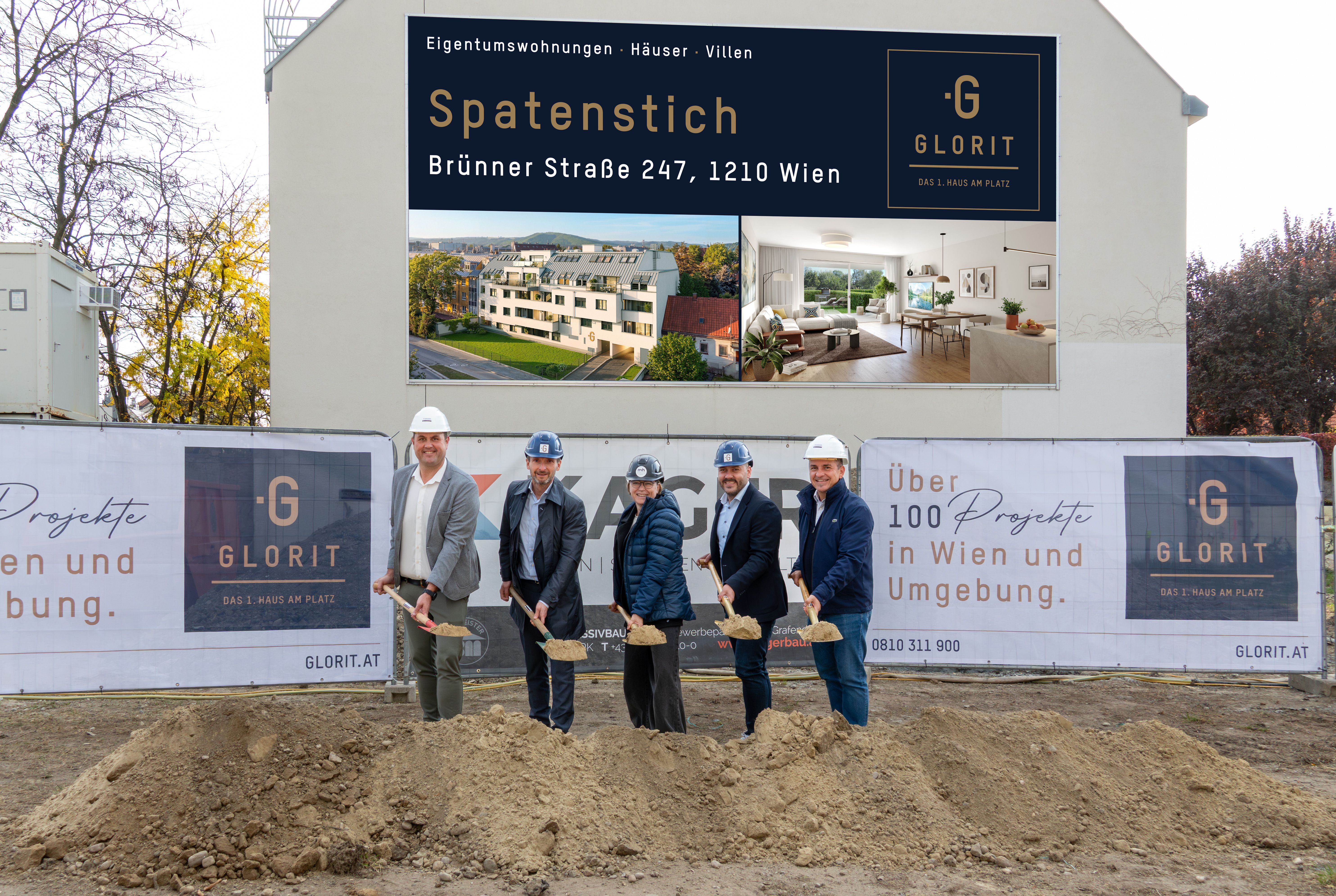Fünf Personen mit Helmen heben beim Spatenstich für ein Bauprojekt Erde mit Schaufeln aus.