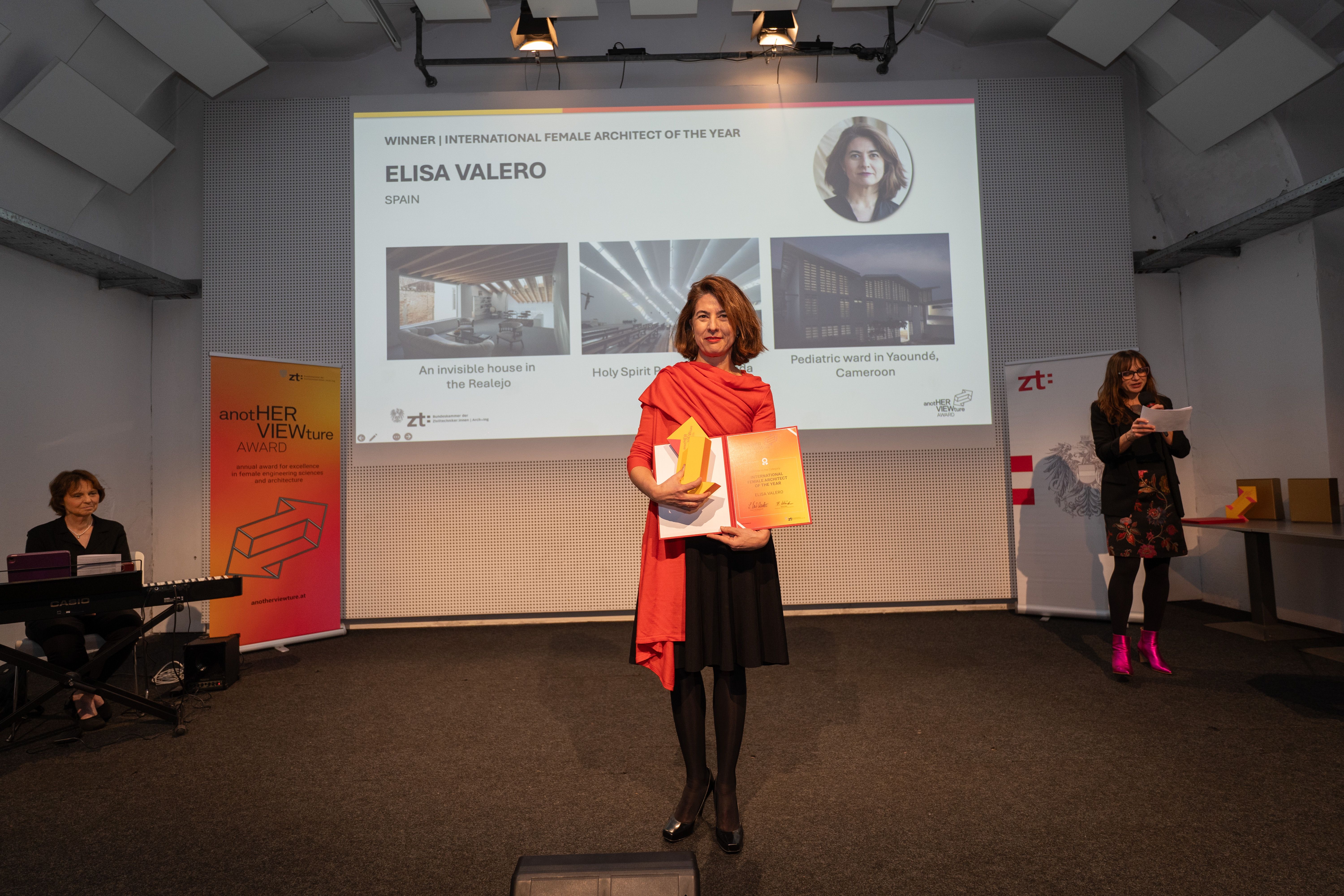 Frau erhält eine Auszeichnung auf einer Bühne, im Hintergrund eine Präsentation über Elisa Valero.