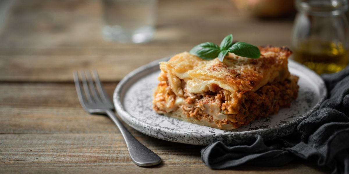 Ein Stück Lasagne mit Basilikum auf einem Teller, daneben eine Gabel auf Holztisch.