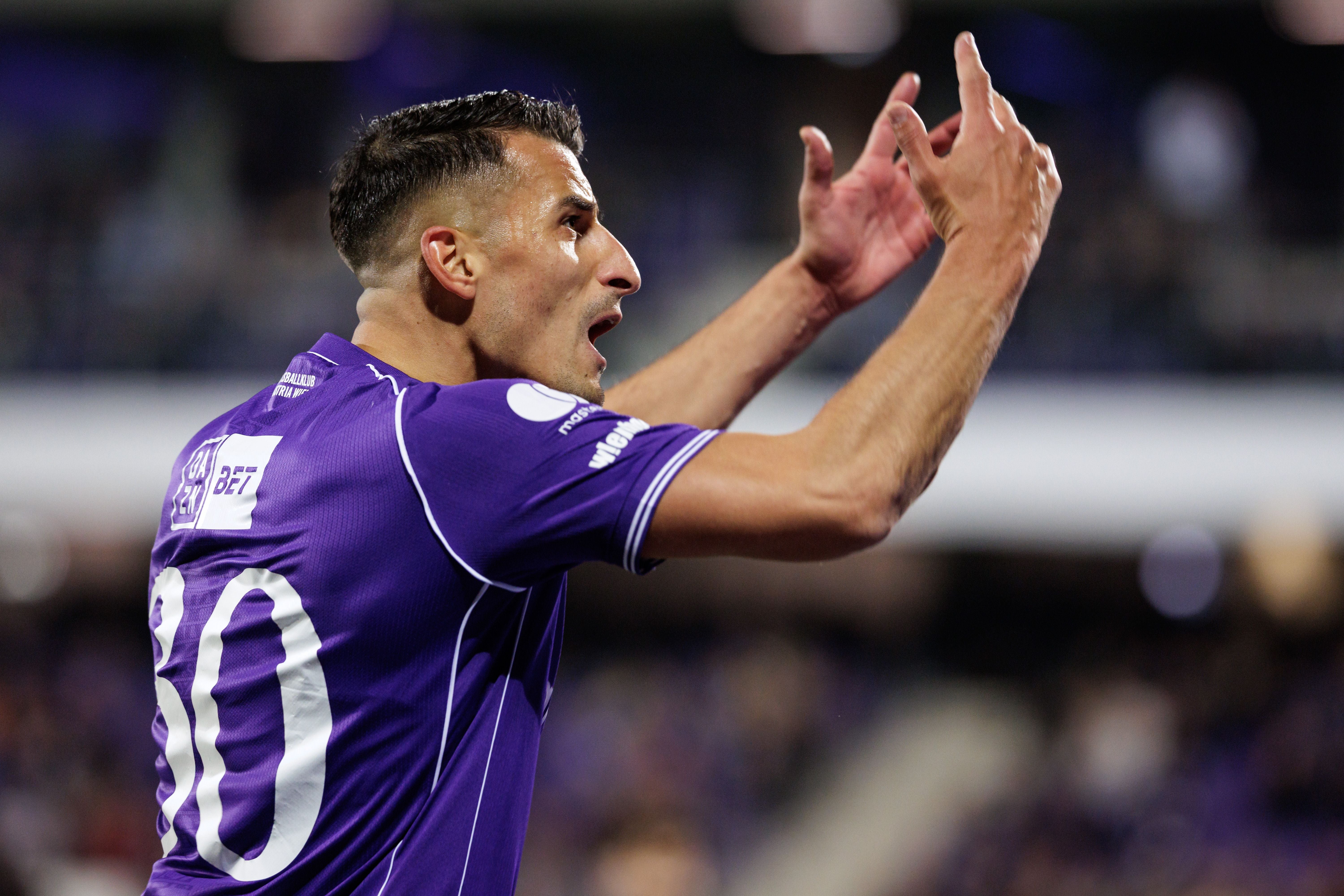Ein Fußballspieler im violetten Trikot gestikuliert mit erhobenen Händen auf dem Spielfeld.