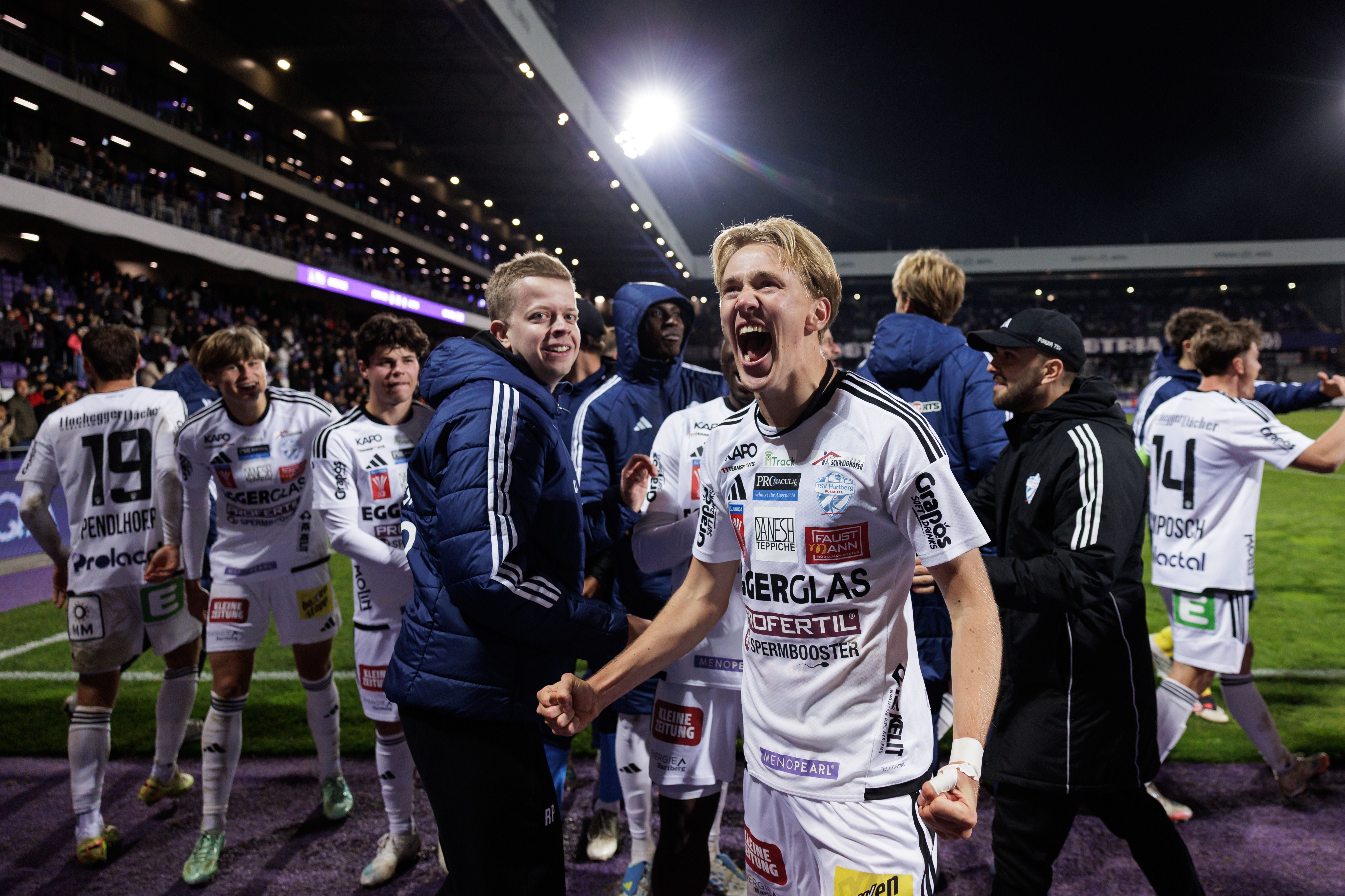 Jubelnde Hartberg-Spieler feiern einen Sieg auf dem Fußballfeld bei Nacht.