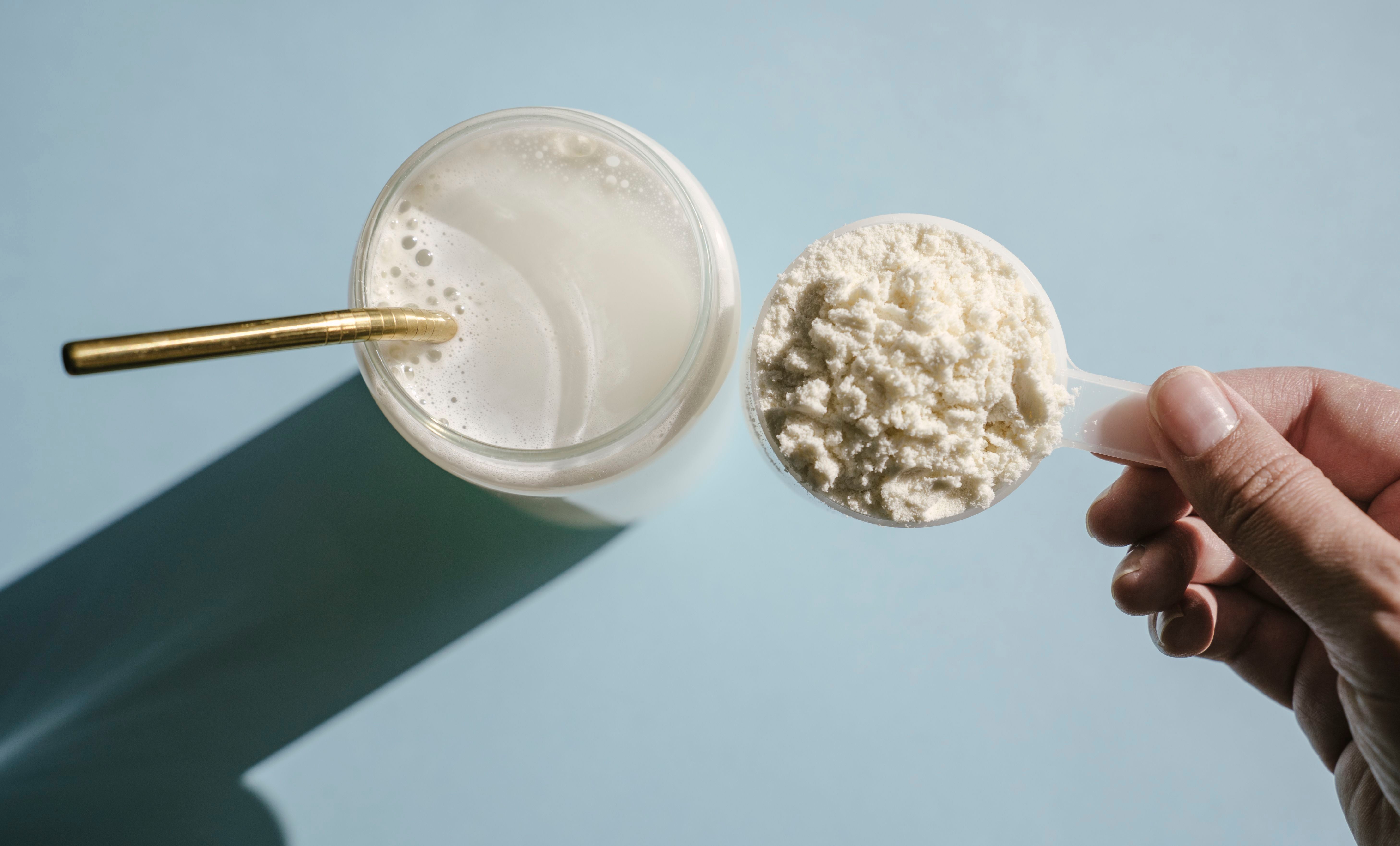Messlöffel mit Proteinpulver neben einem Glas Proteinshake mit Strohhalm auf hellem Untergrund.