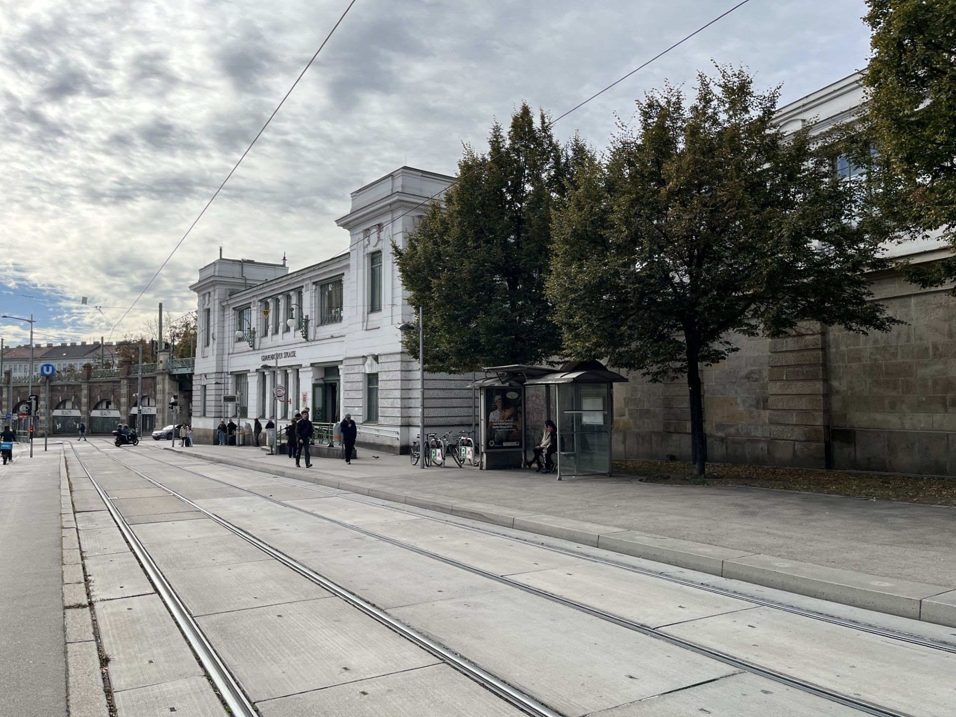 Straßenbahngleise vor einer weißen U-Bahn-Station mit Bäumen und wartenden Personen an der Haltestelle.