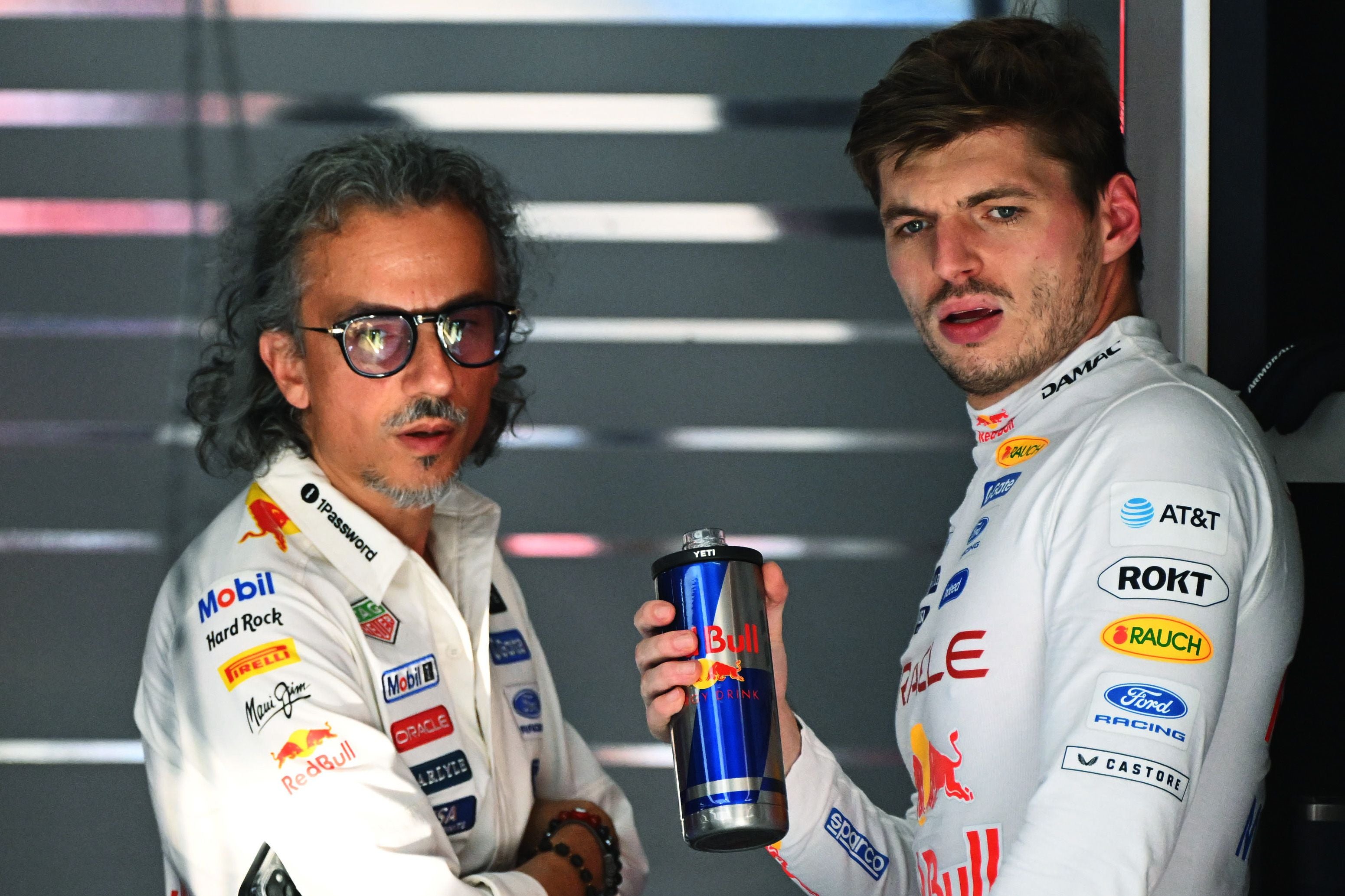 Zwei Männer im Red Bull Racing Teamoutfit sprechen in einer Boxengasse, einer hält eine Red Bull-Dose.