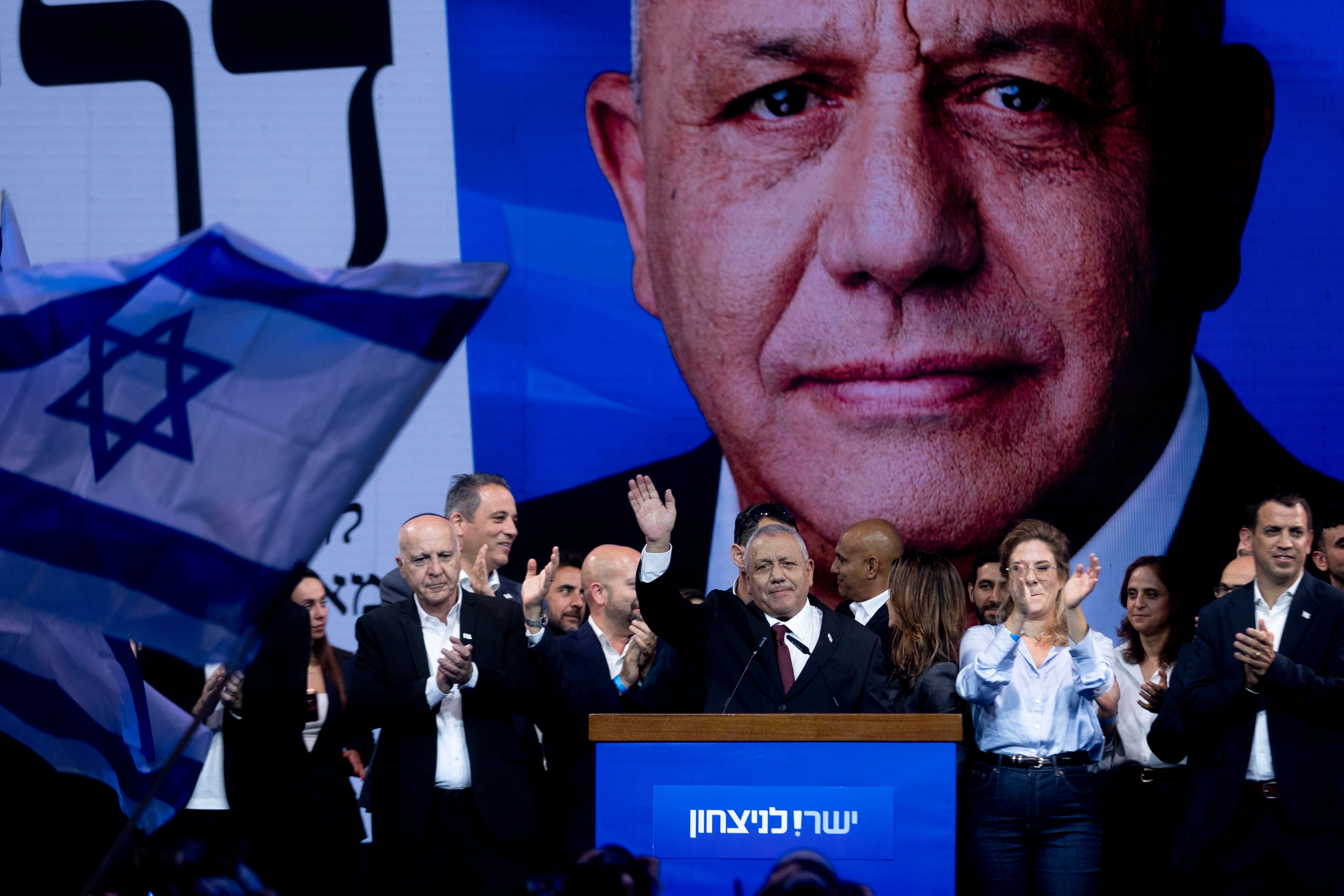 Ein Politiker steht auf einer Bühne und winkt, umgeben von applaudierenden Menschen und einer israelischen Fahne.