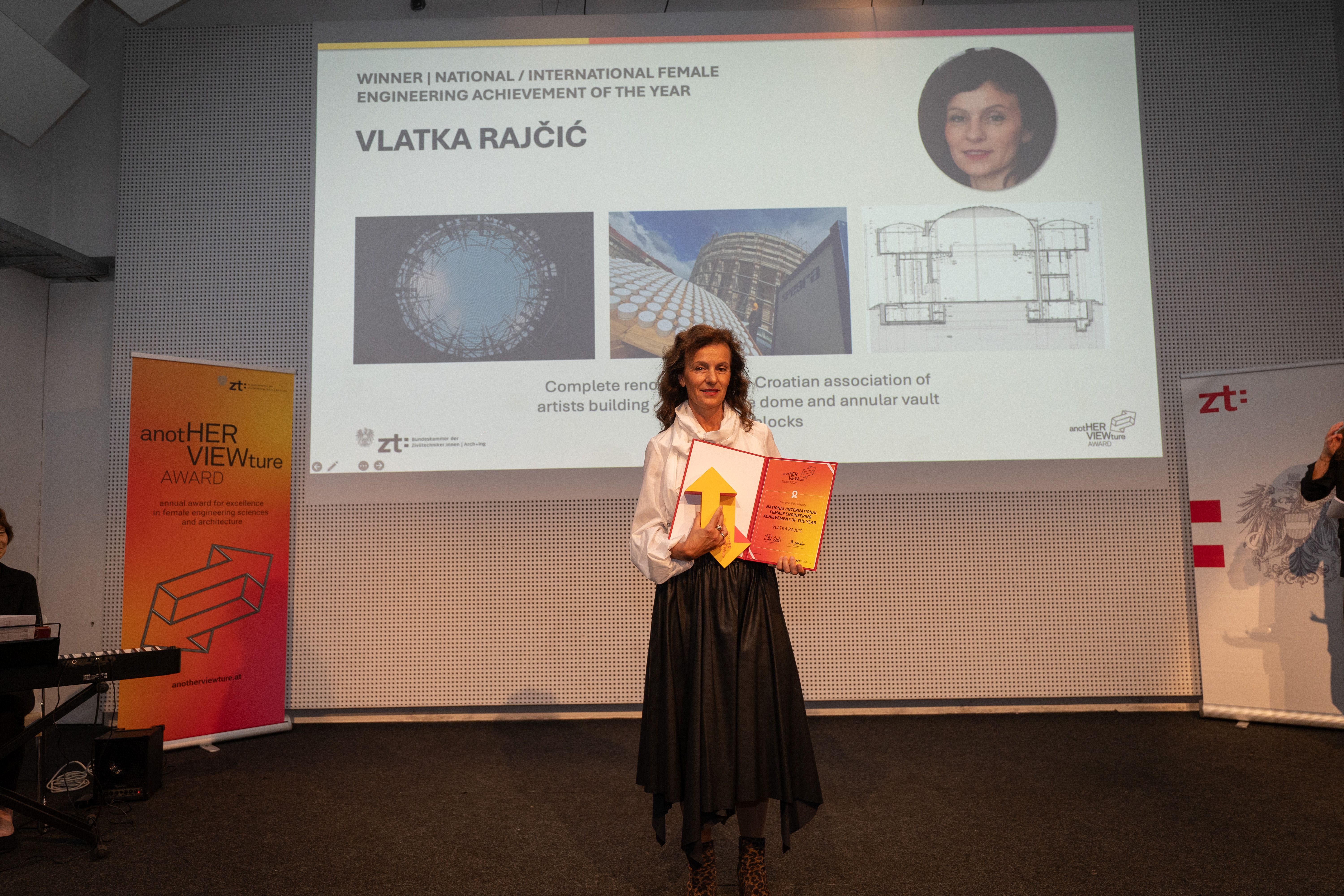 Frau mit Auszeichnung steht auf einer Bühne vor einer Präsentationsfolie mit ihrem Namen und Foto.