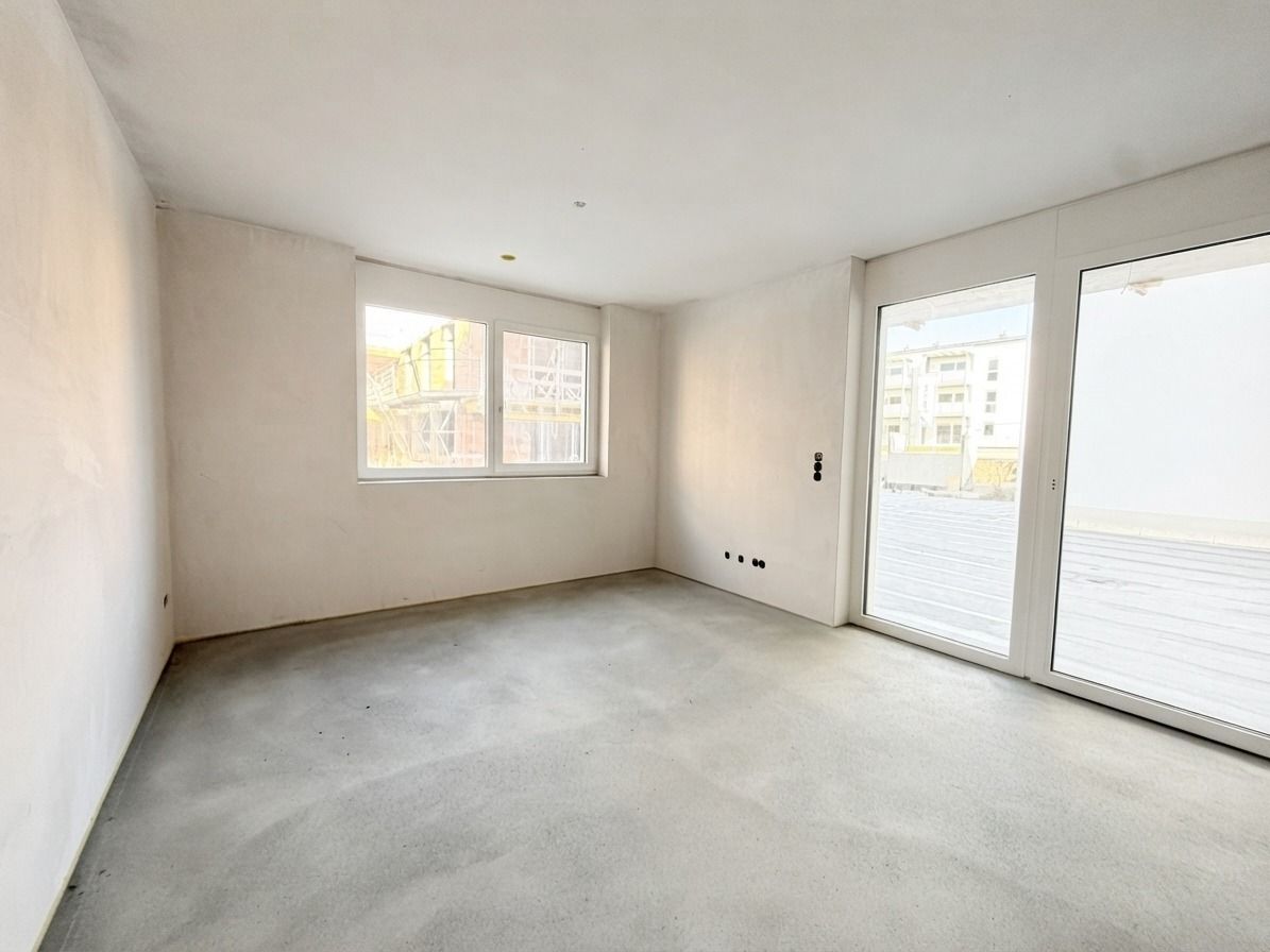 Leerer, heller Raum mit Betonboden, weißen Wänden und großen Fenstern mit Blick nach draußen.
