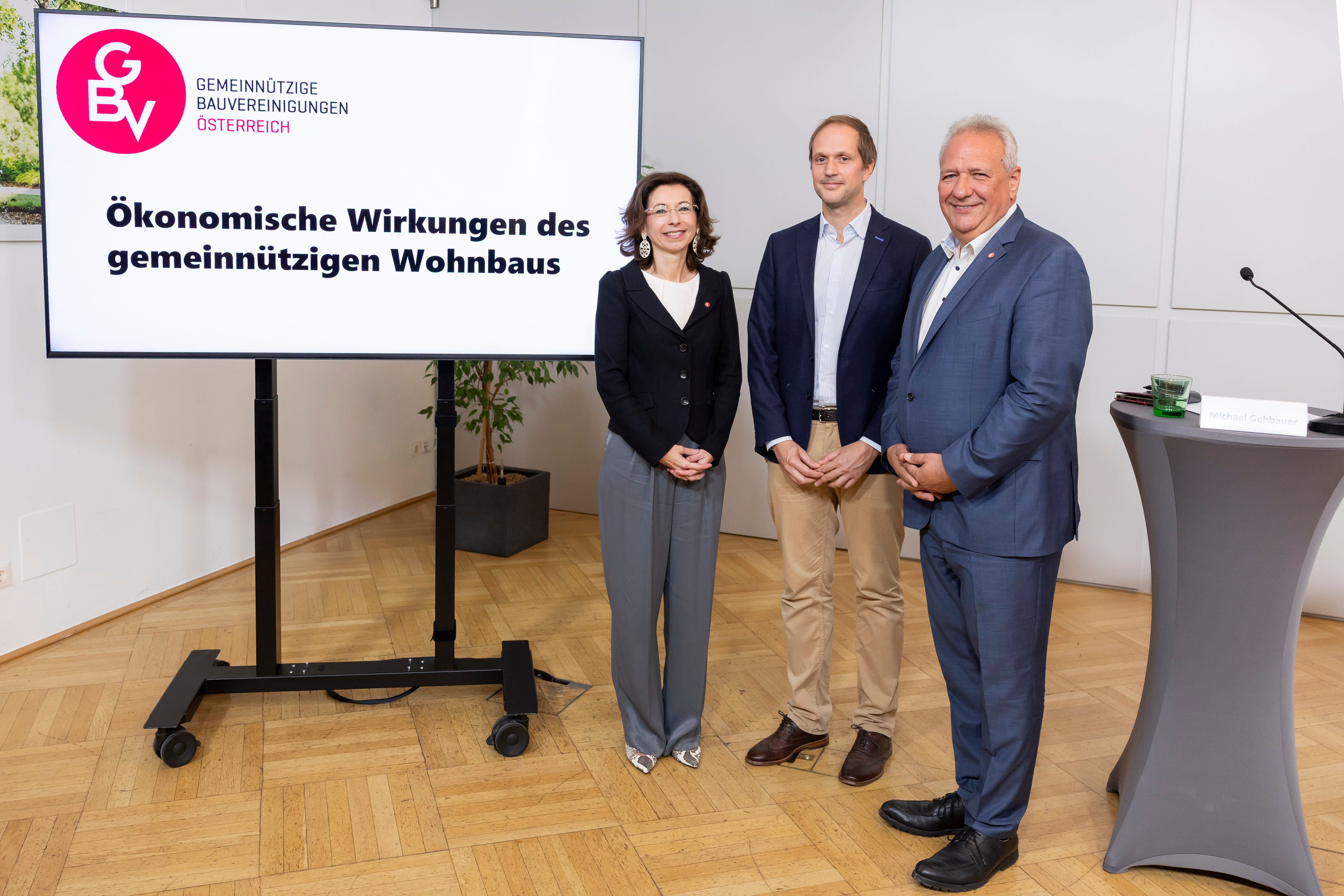 Drei Personen stehen neben einem Bildschirm mit der Aufschrift „Ökonomische Wirkungen des gemeinnützigen Wohnbaus“.