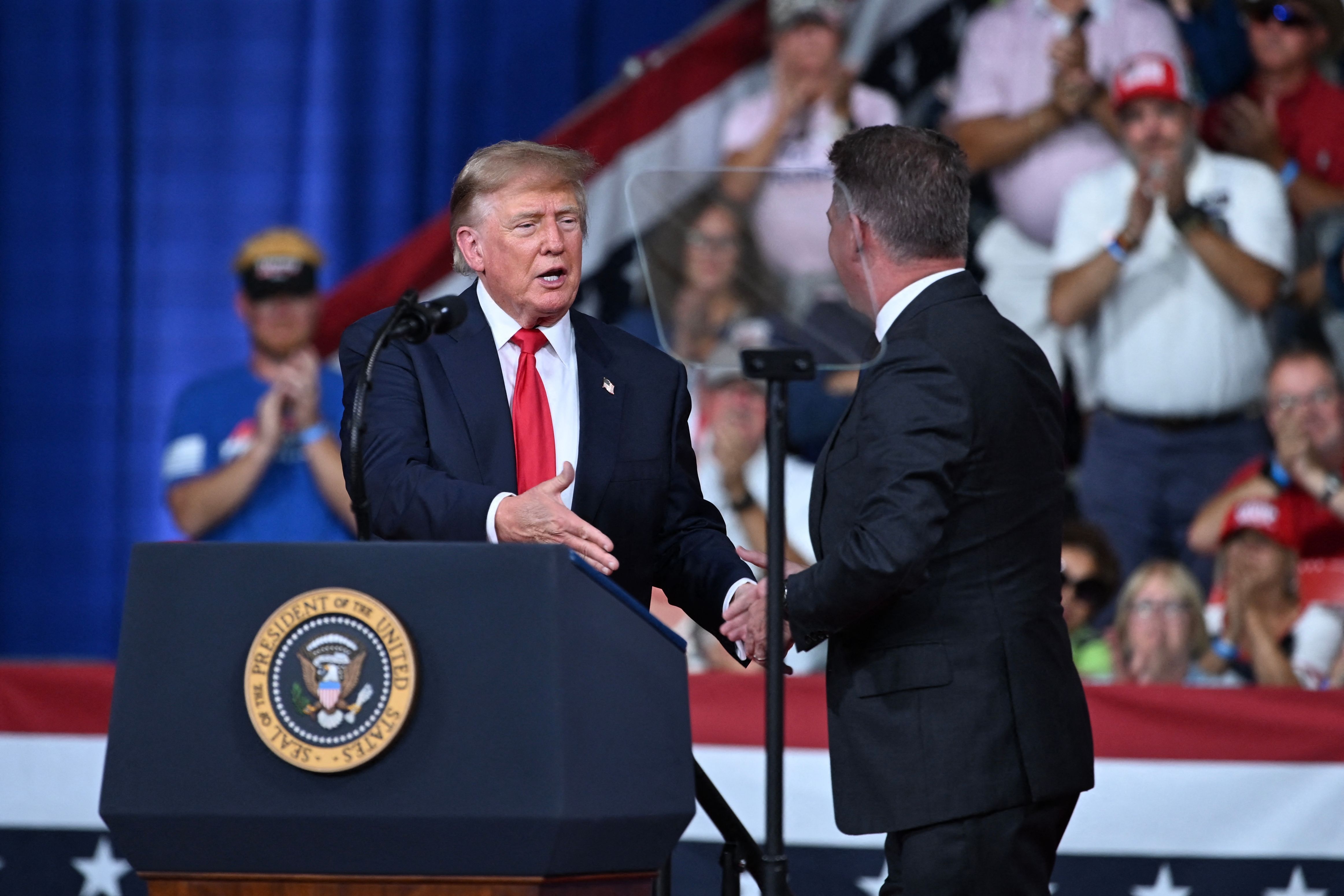 Donald Trump begrüßt einen Mann auf einer Bühne bei einer Wahlkampfveranstaltung in einer vollen Halle.
