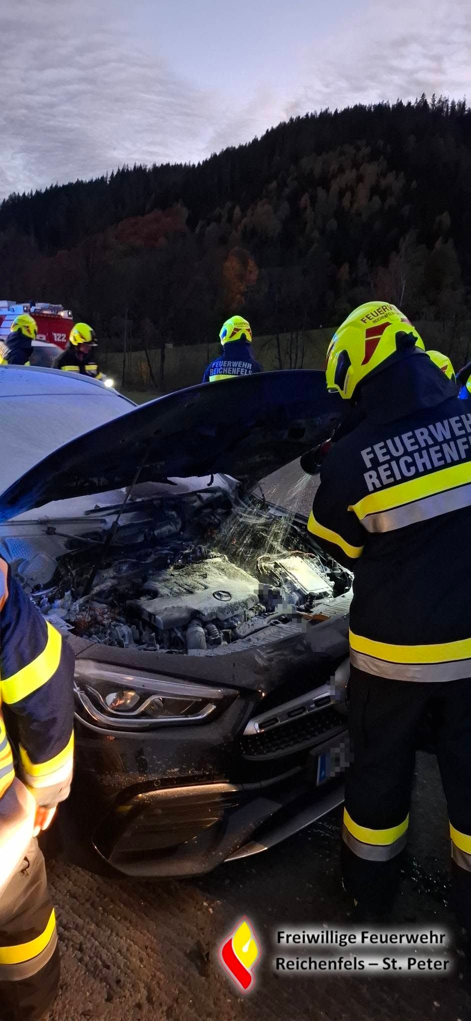 Feuerwehr löscht einen brennenden Motorraum eines Autos im Freien bei Dämmerung.