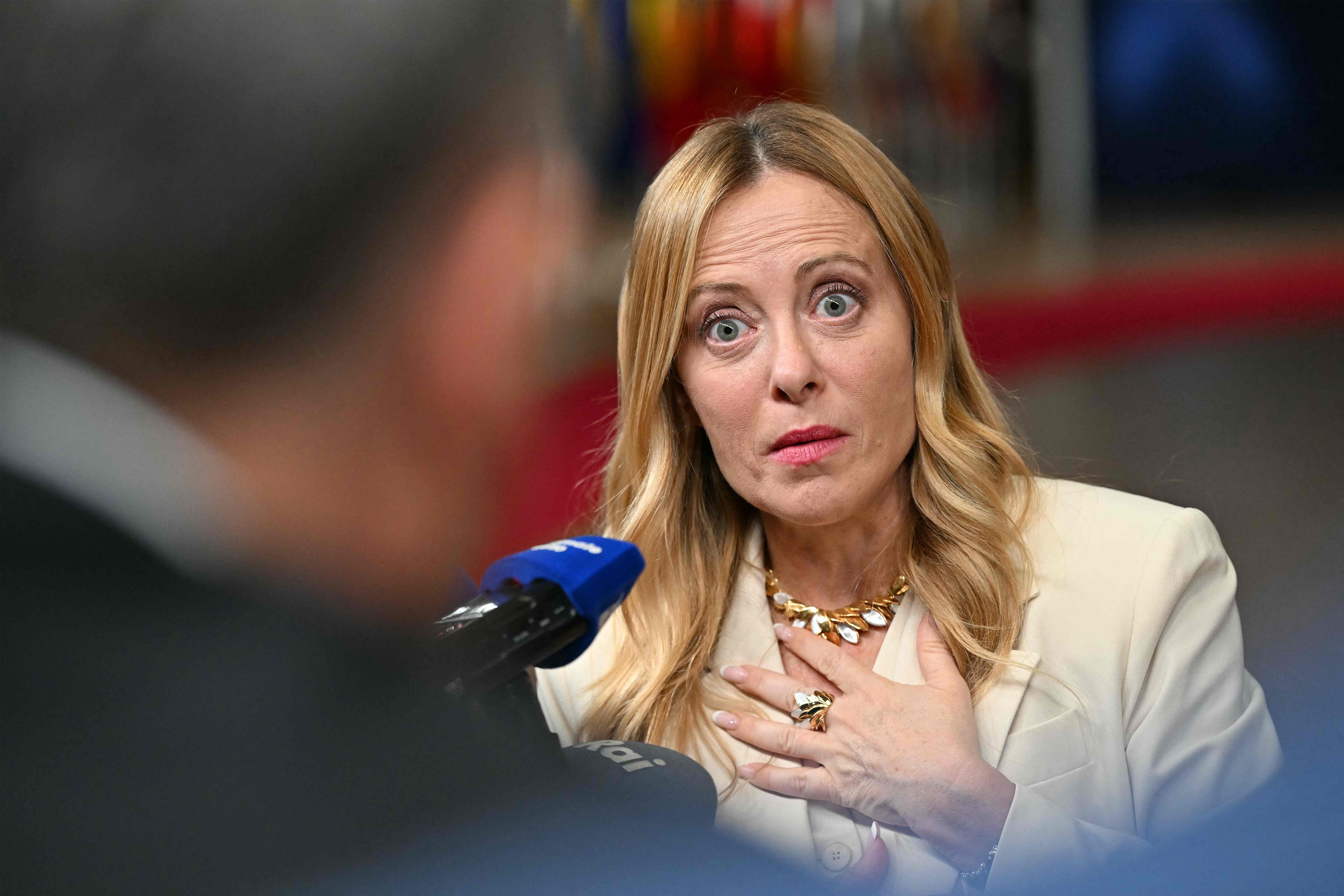 Eine Frau mit blondem Haar spricht, wirkt überrascht und hält die Hand auf die Brust.