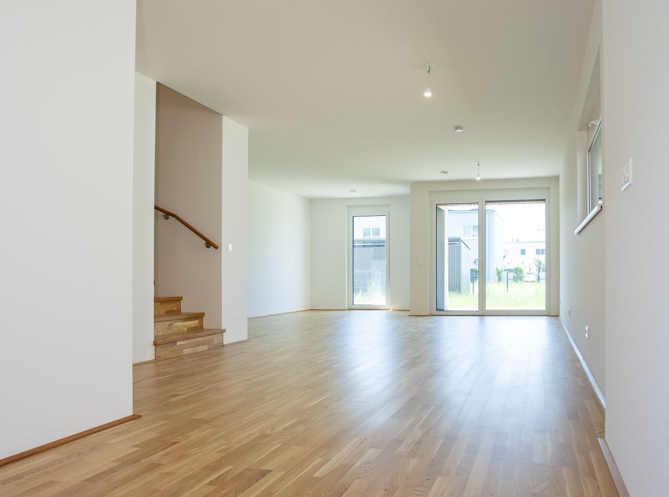 Helles, leeres Wohnzimmer mit Parkettboden, Treppe und großen Fenstern zum Garten.