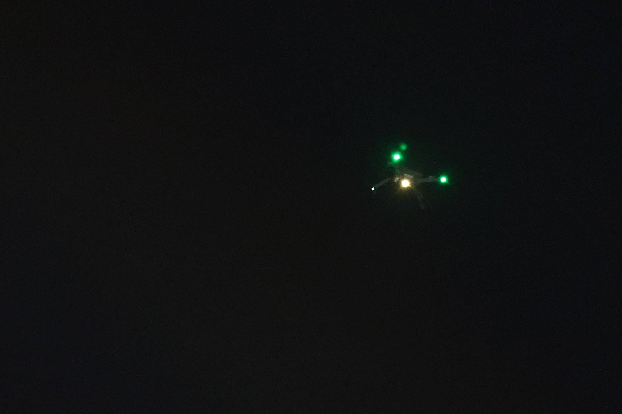 Eine Drohne mit grünen Lichtern fliegt nachts über einem dunklen Himmel.