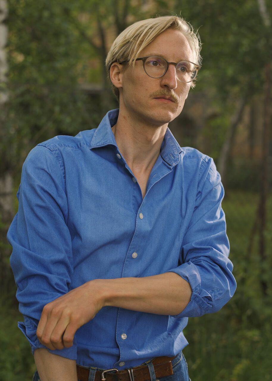 Mann mit blonden Haaren und Brille trägt blaues Hemd und steht im Freien vor Bäumen.