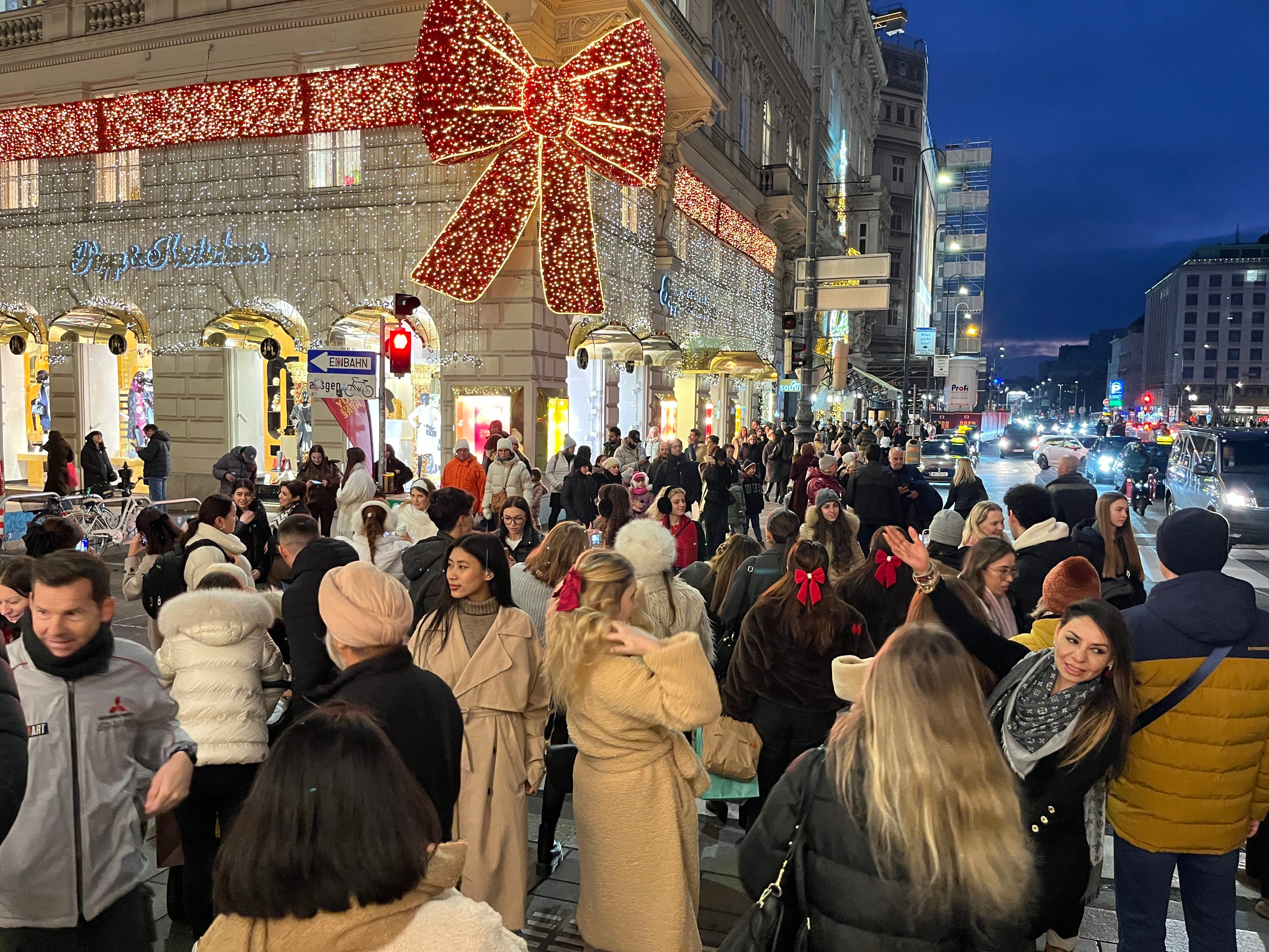 Viele Menschen spazieren abends durch eine festlich beleuchtete Einkaufsstraße mit XXL-Weihnachtsschleife.