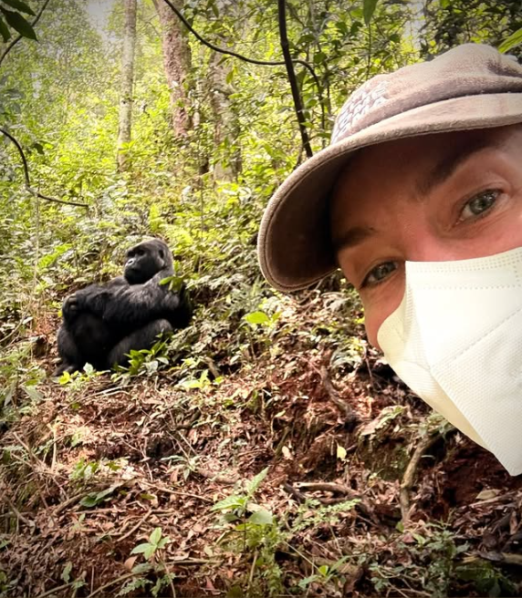 Person mit Maske im Wald, im Hintergrund sitzt ein Gorilla im dichten Grün.