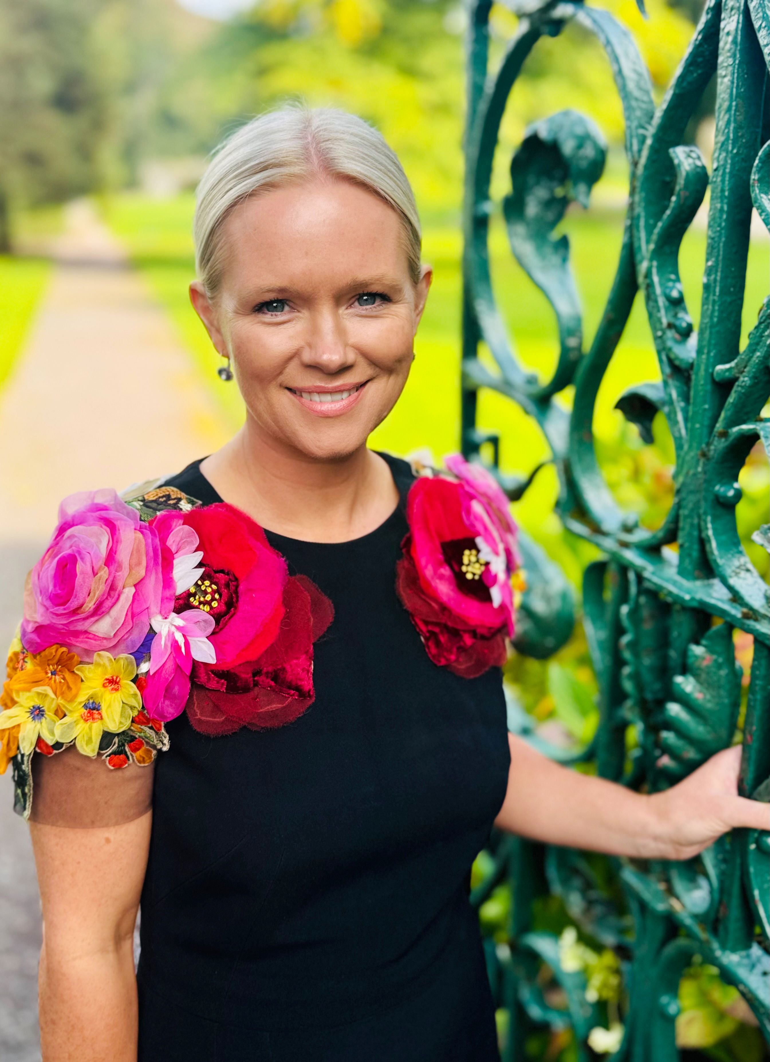 Frau mit Blumenapplikationen auf den Schultern steht lächelnd an einem grünen Zaun im Garten.