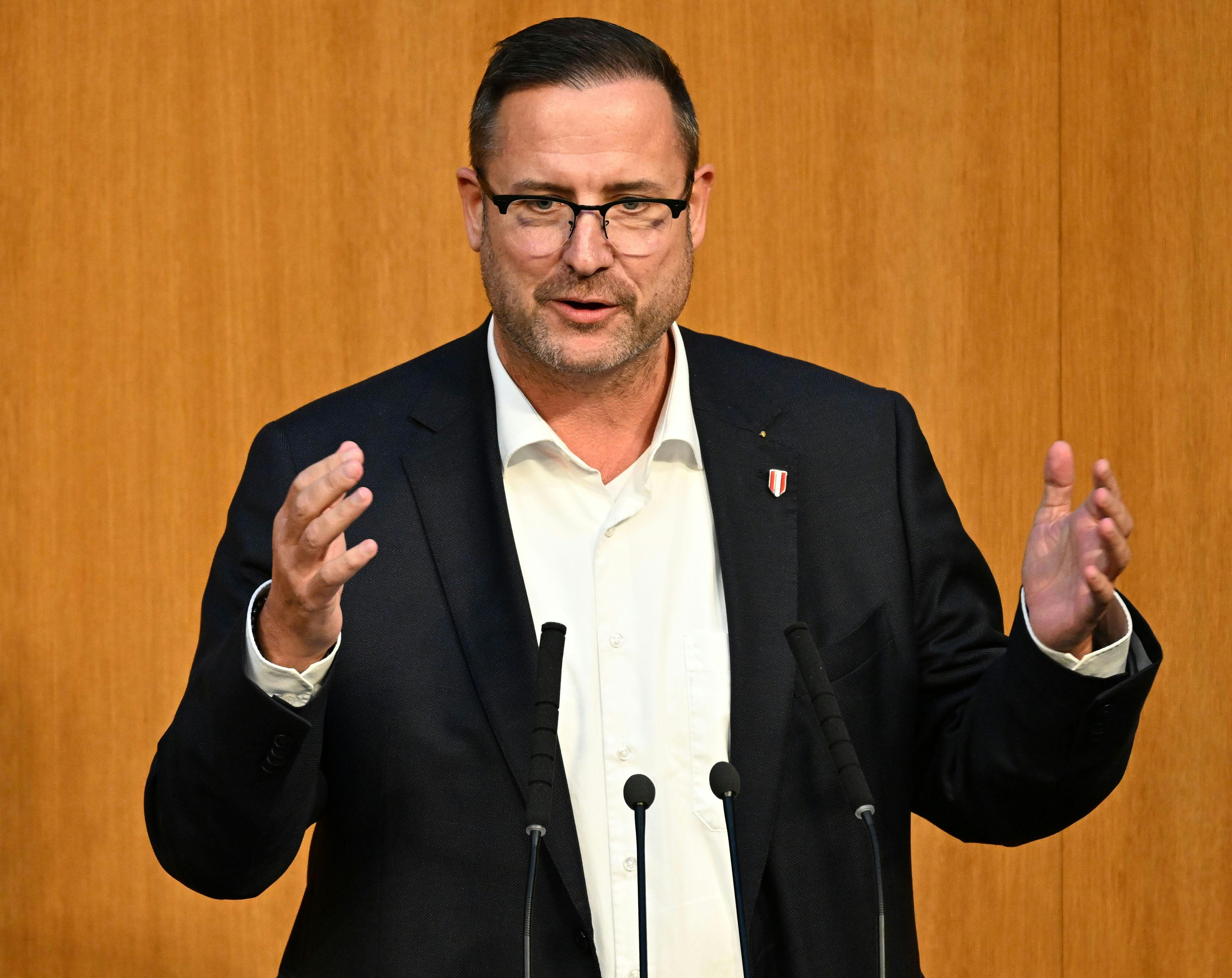Ein Mann in Anzug und Brille spricht gestikulierend am Rednerpult im Parlament.