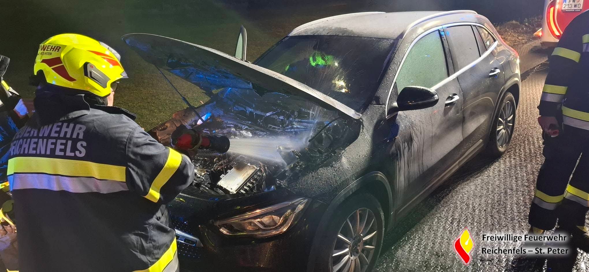 Feuerwehr löscht brennenden Motorraum eines schwarzen Autos auf einer Straße bei Nacht.