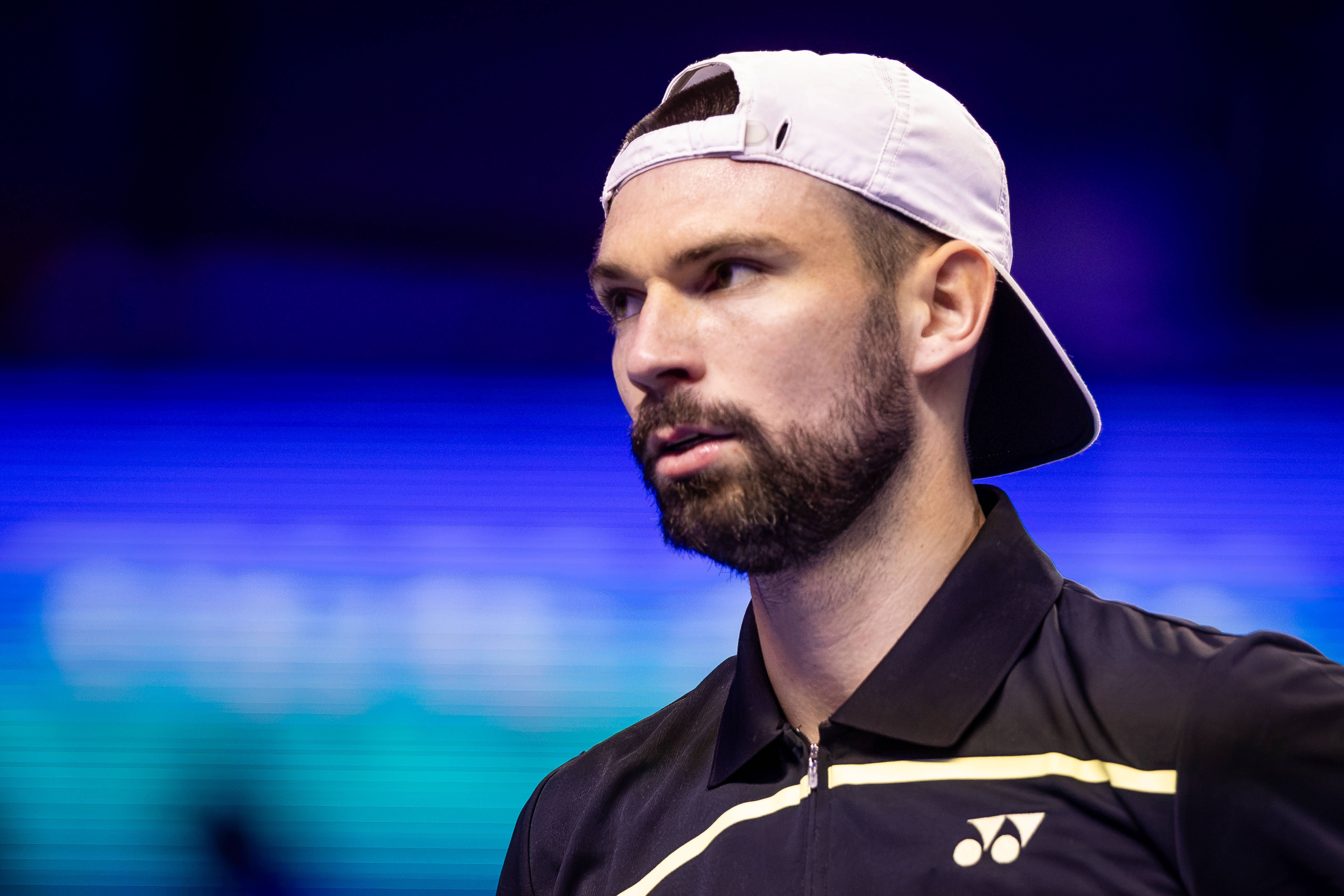 Ein Tennisspieler mit Bart und Mütze rückwärts trägt ein schwarzes Shirt bei einem Turnier.