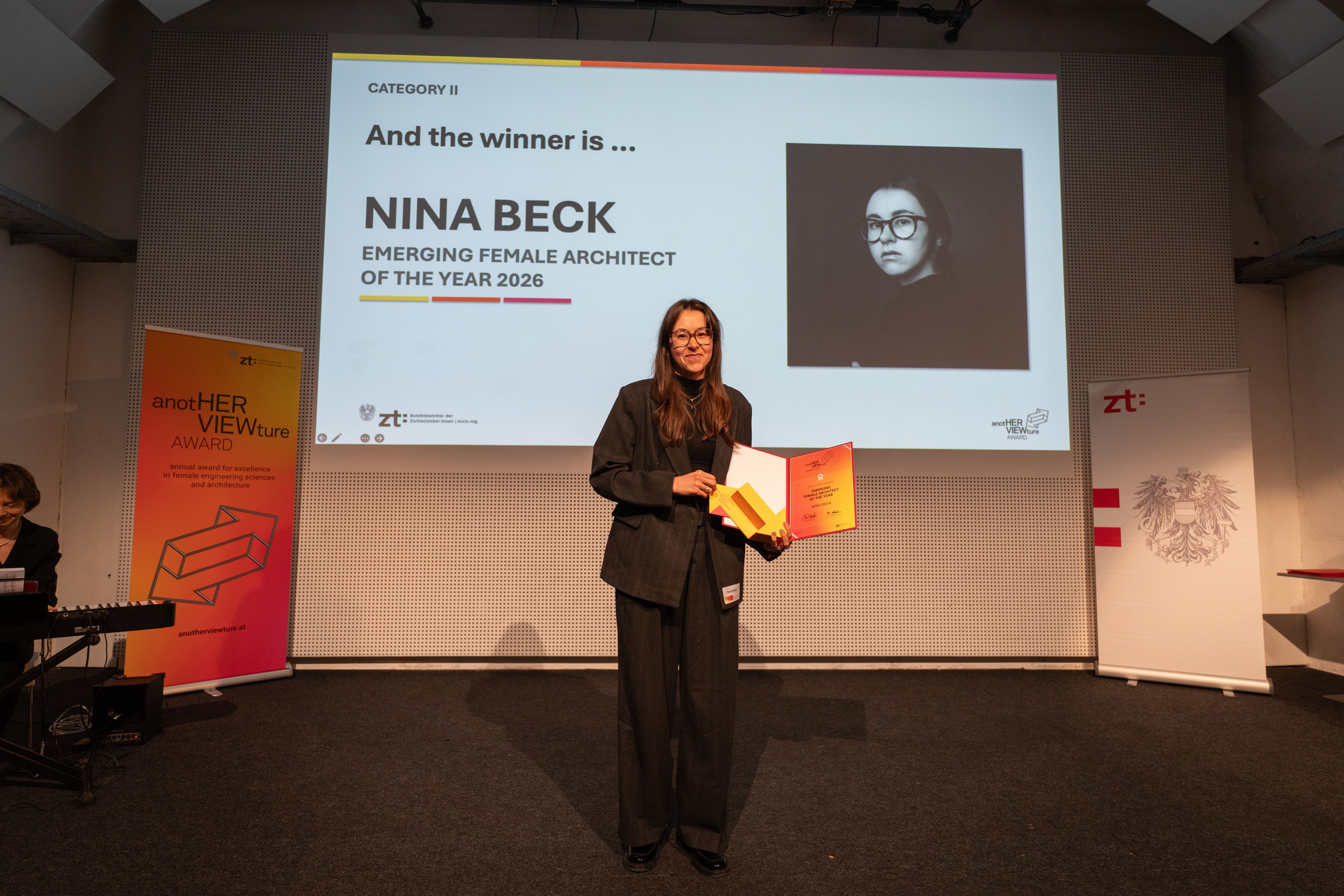 Frau mit Urkunde steht vor Leinwand, die sie als Preisträgerin in Architektur auszeichnet.