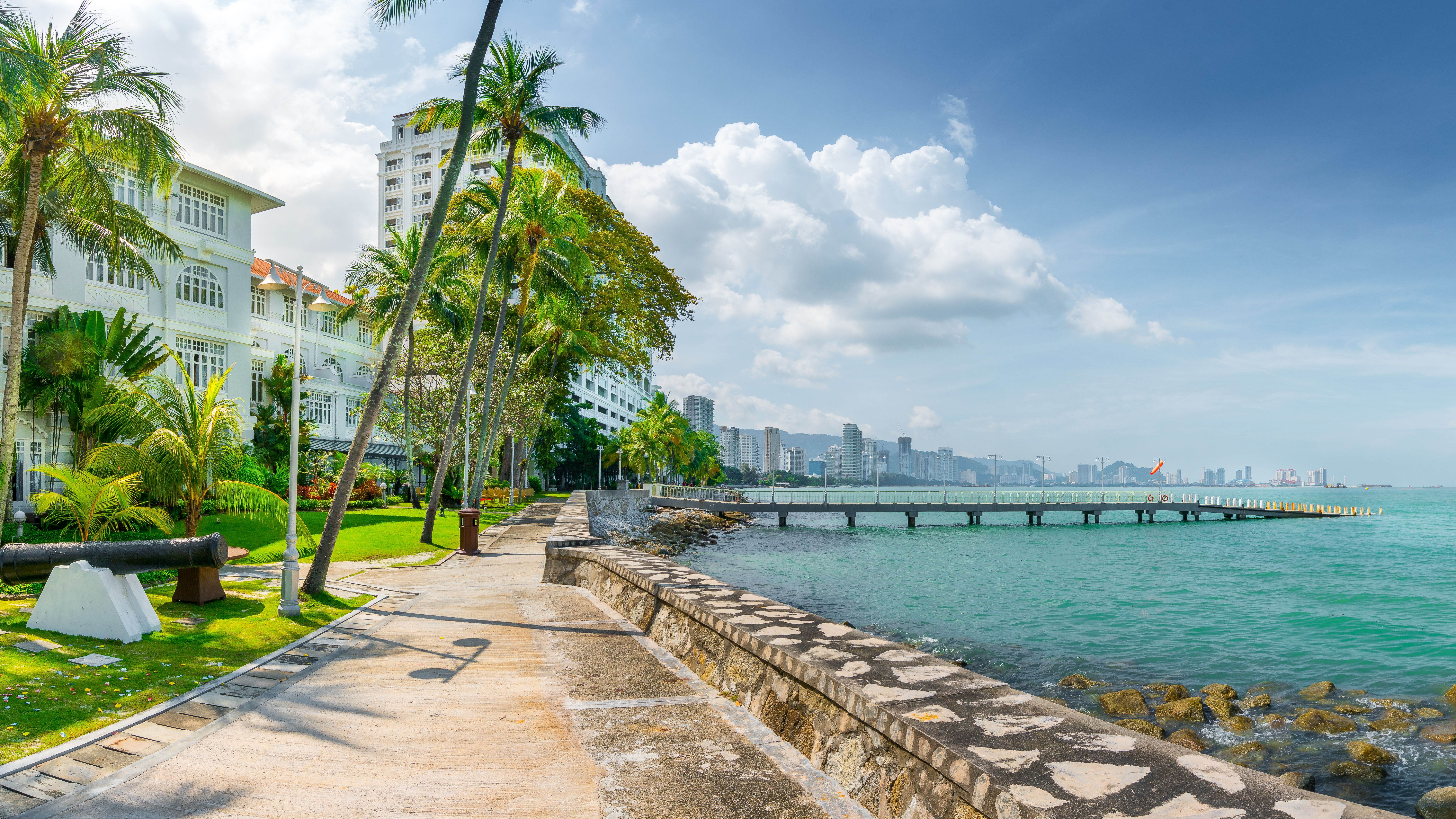 Promenade mit Palmen am Meer in Penang, Malaysia, mit Blick auf Hochhäuser und Steg.