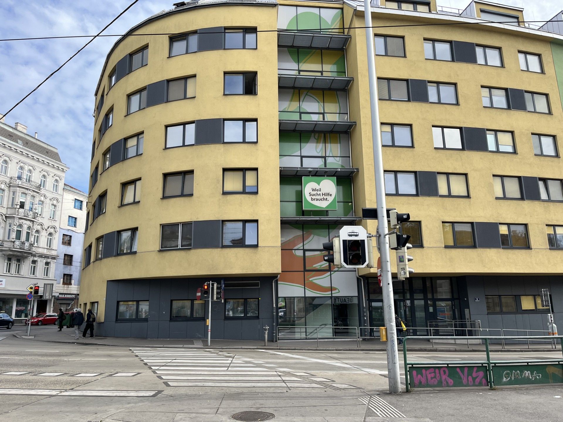 Gelbes Eckgebäude an einer Kreuzung mit einem „Well. Sucht Hilfe braucht.“-Schild.