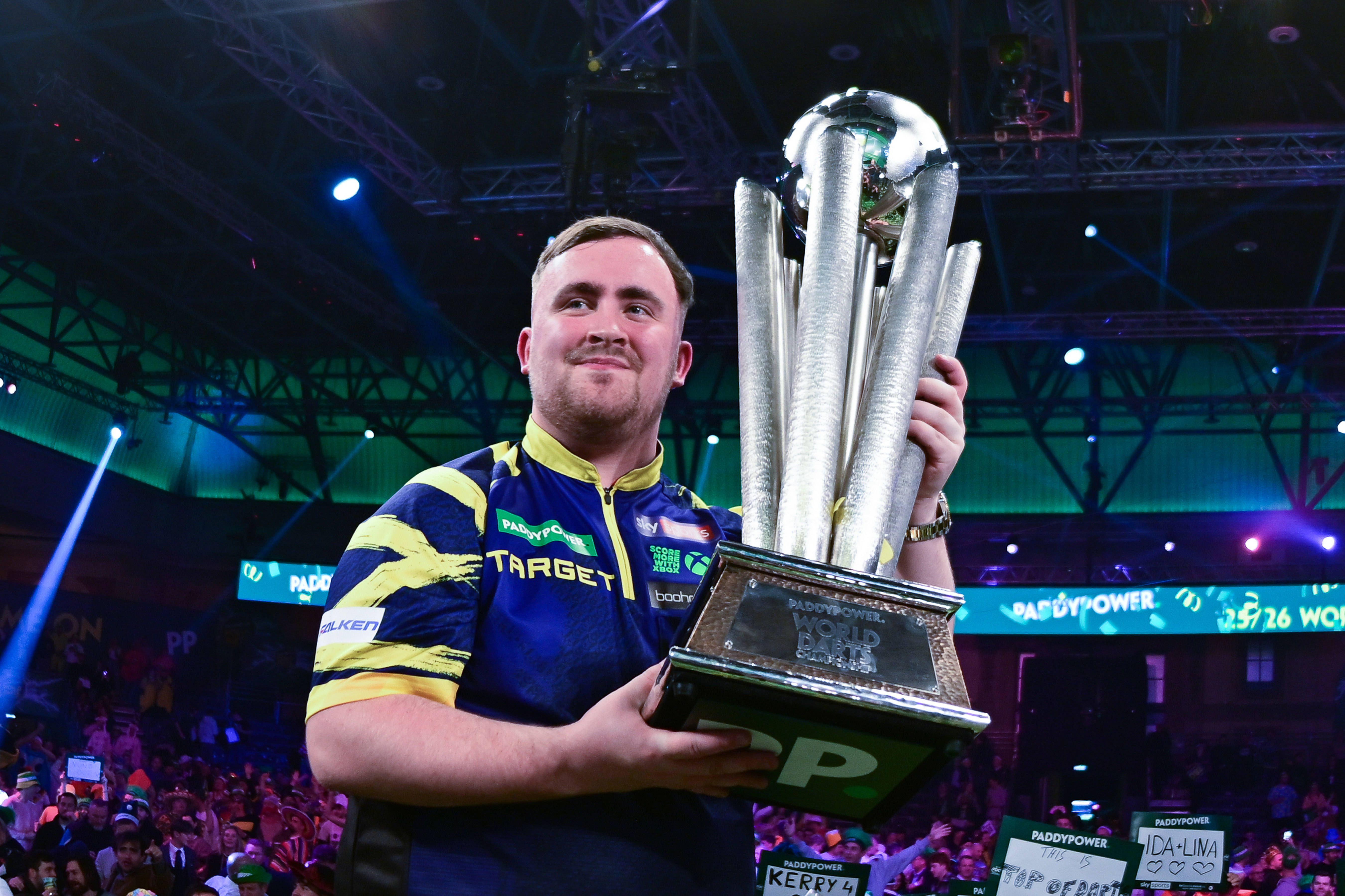 Ein junger Mann hält stolz einen großen Pokal bei einer Dartmeisterschaft in einer Arena hoch.